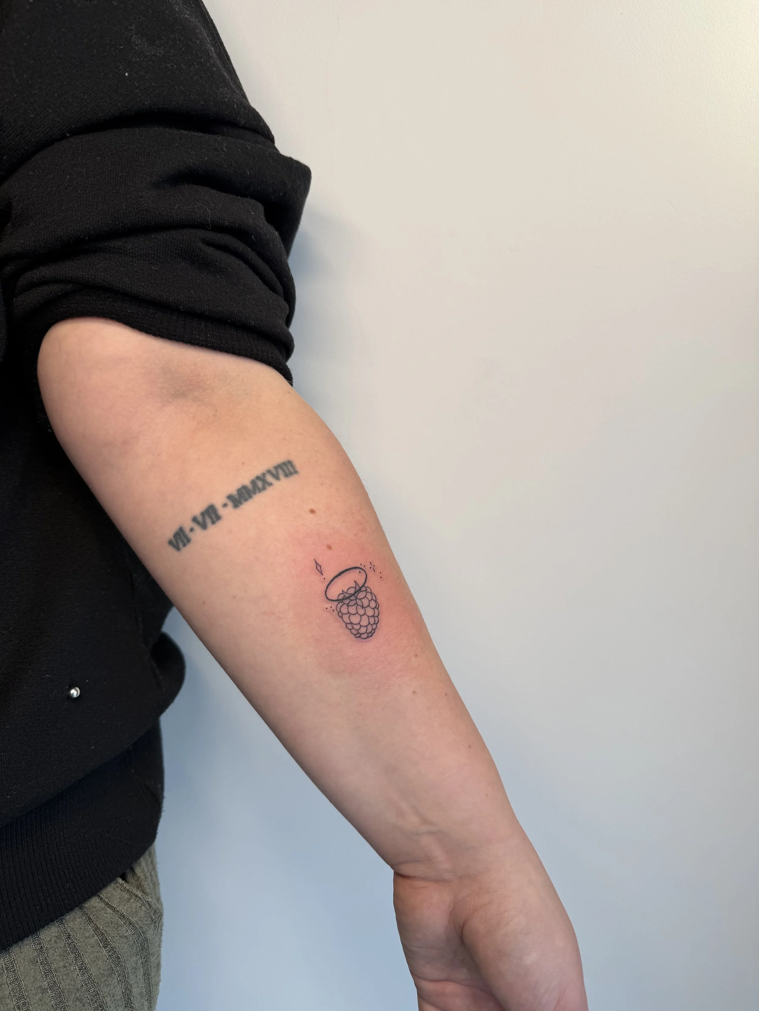 De afbeelding toont een close-up van een onderarm met een tatoeage van een bosbessenblad en een paar bessen. De persoon draagt een zwarte trui met gerolde mouwen en een lichte broek met strepen.