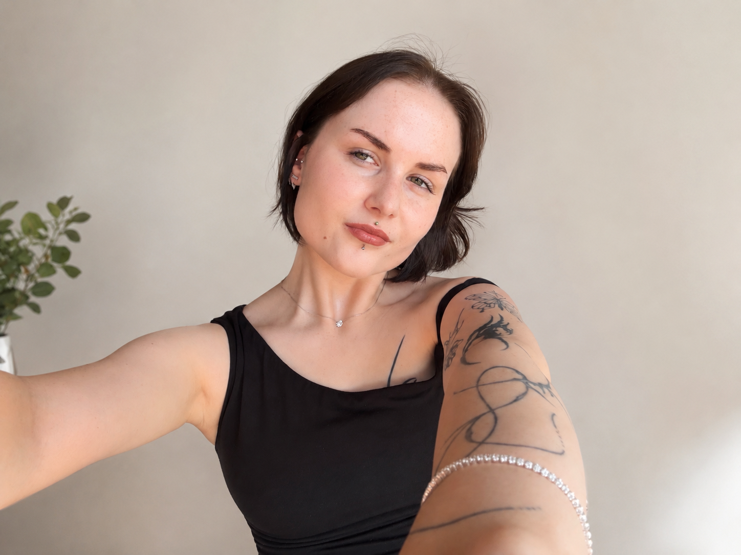 Een jonge vrouw met korte bruin haar, die een selfie neemt. Ze draagt een zwart topje, sieraden en heeft tatoeages op haar arm. Ze staat voor een lichte muur met een plant op de achtergrond.
