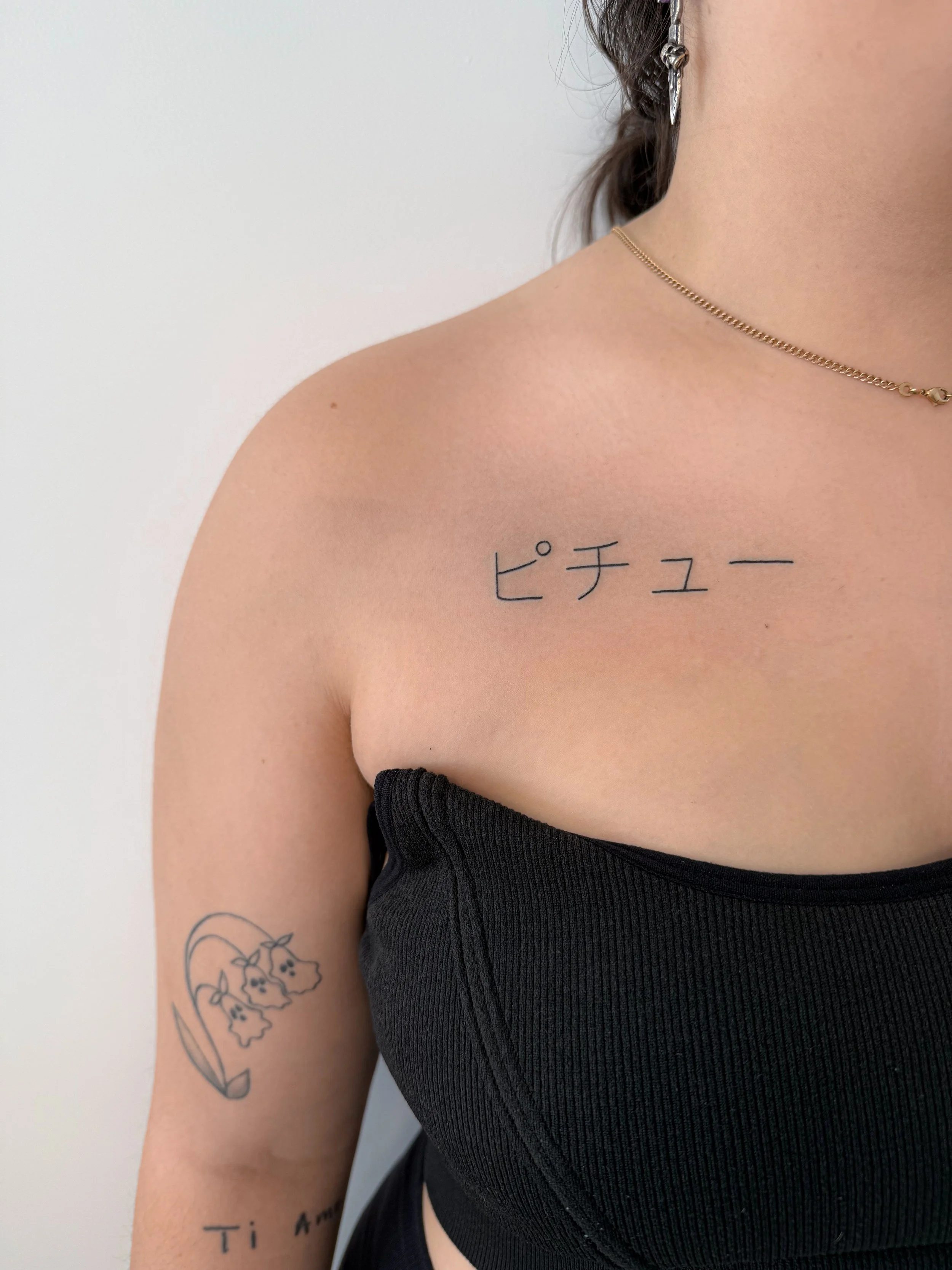 Close-up van een vrouw met een tatoeage van bloemen op haar linkermouw en een tekst onder haar arm, ze draagt een gouden ketting en een oorbel, gekleed in een zwart ribbelachtig Topje.