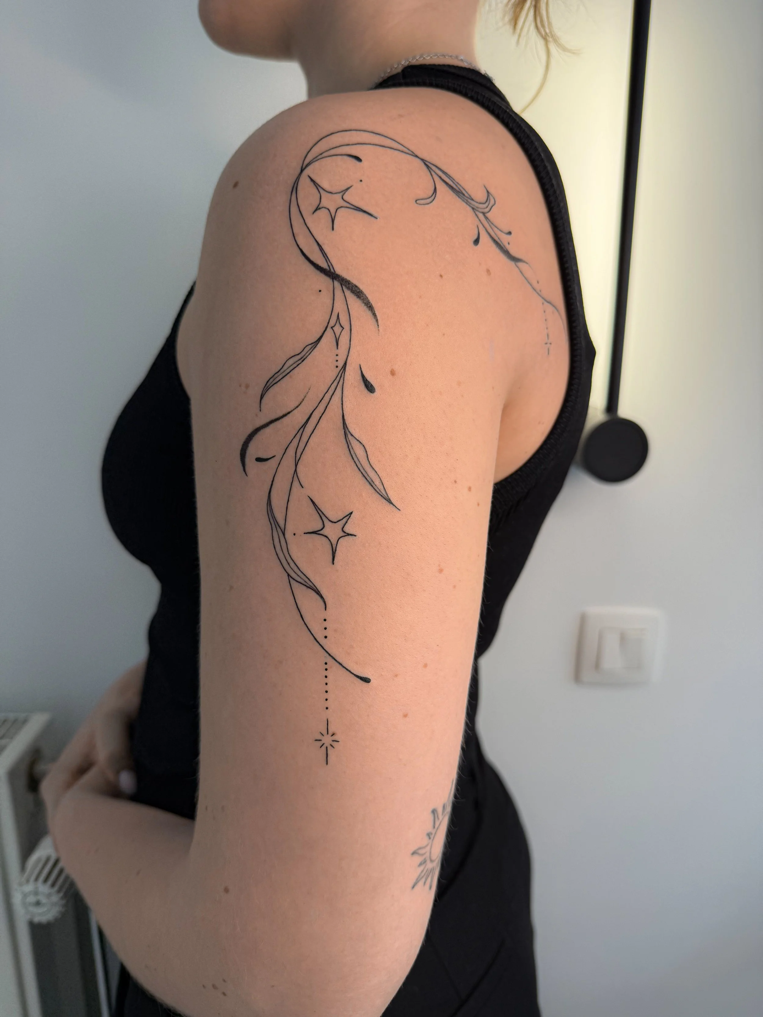 Close-up van een persoon met een bloemen- en sterretjestas a onarm met dunne zwarte tattoo lijnen, deels zichtbaar in zwart topje, achtergrond met witte muur en stopcontact.