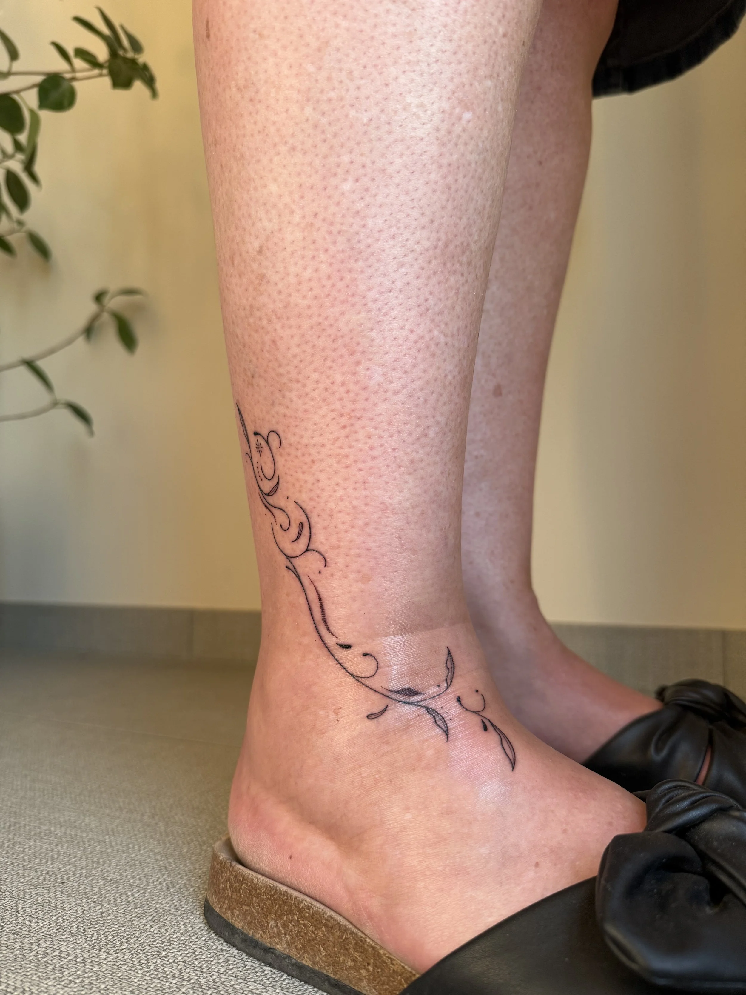 Close-up van een menselijk onderbeen en voet met een schetsachtige tatoeage van een bloem en bloemen op de kuit. De persoon draagt zwarte schoenen en zit op de vloer.