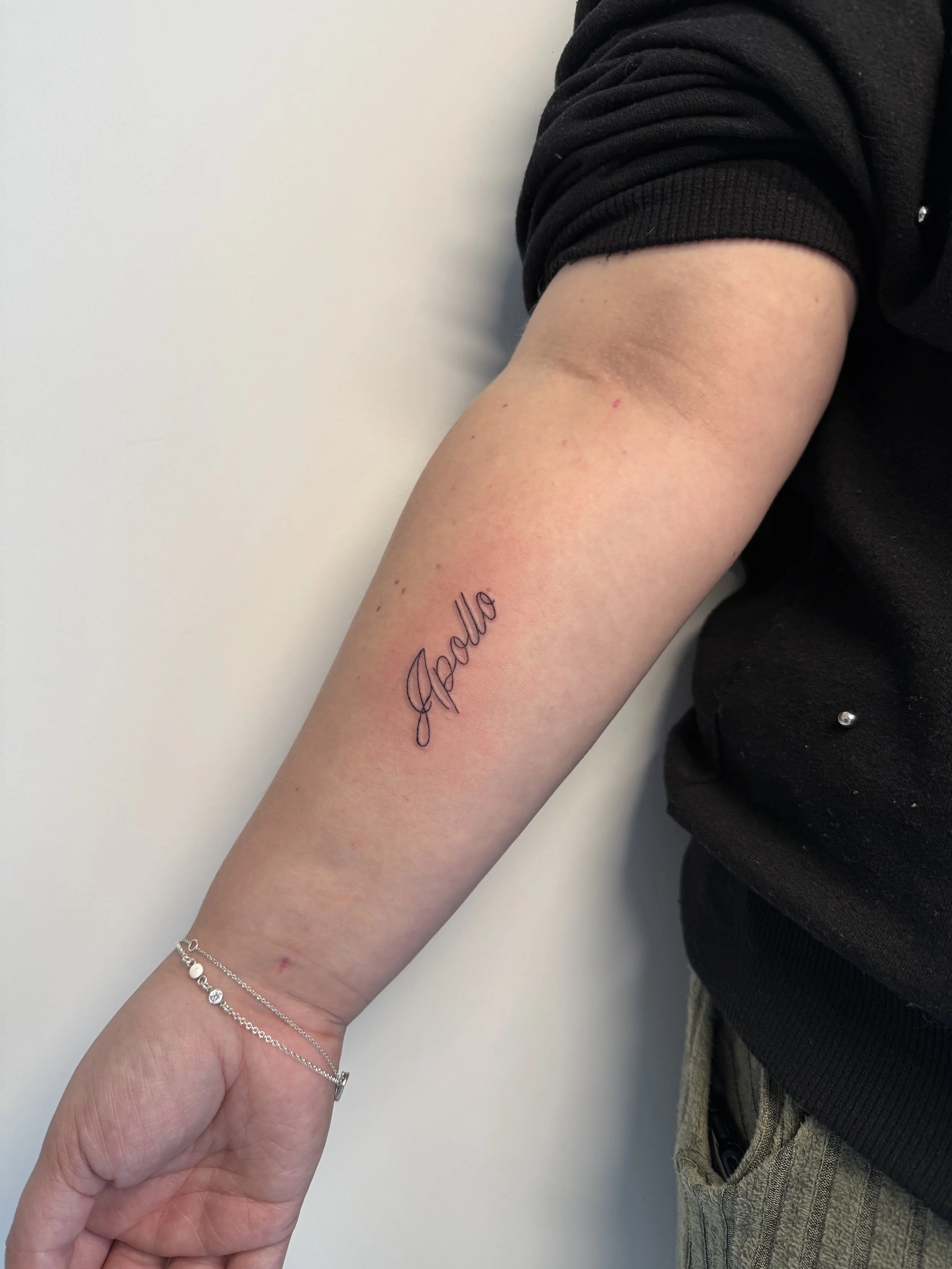 De afbeelding toont een menselijke arm met een tattoo die het woord 'Apollo' schrijft in cursieve letters, een armband met kleine kralen en een dunne ketting. De persoon draagt een zwart kledingstuk en heeft een beetje blootstelling van de taille van