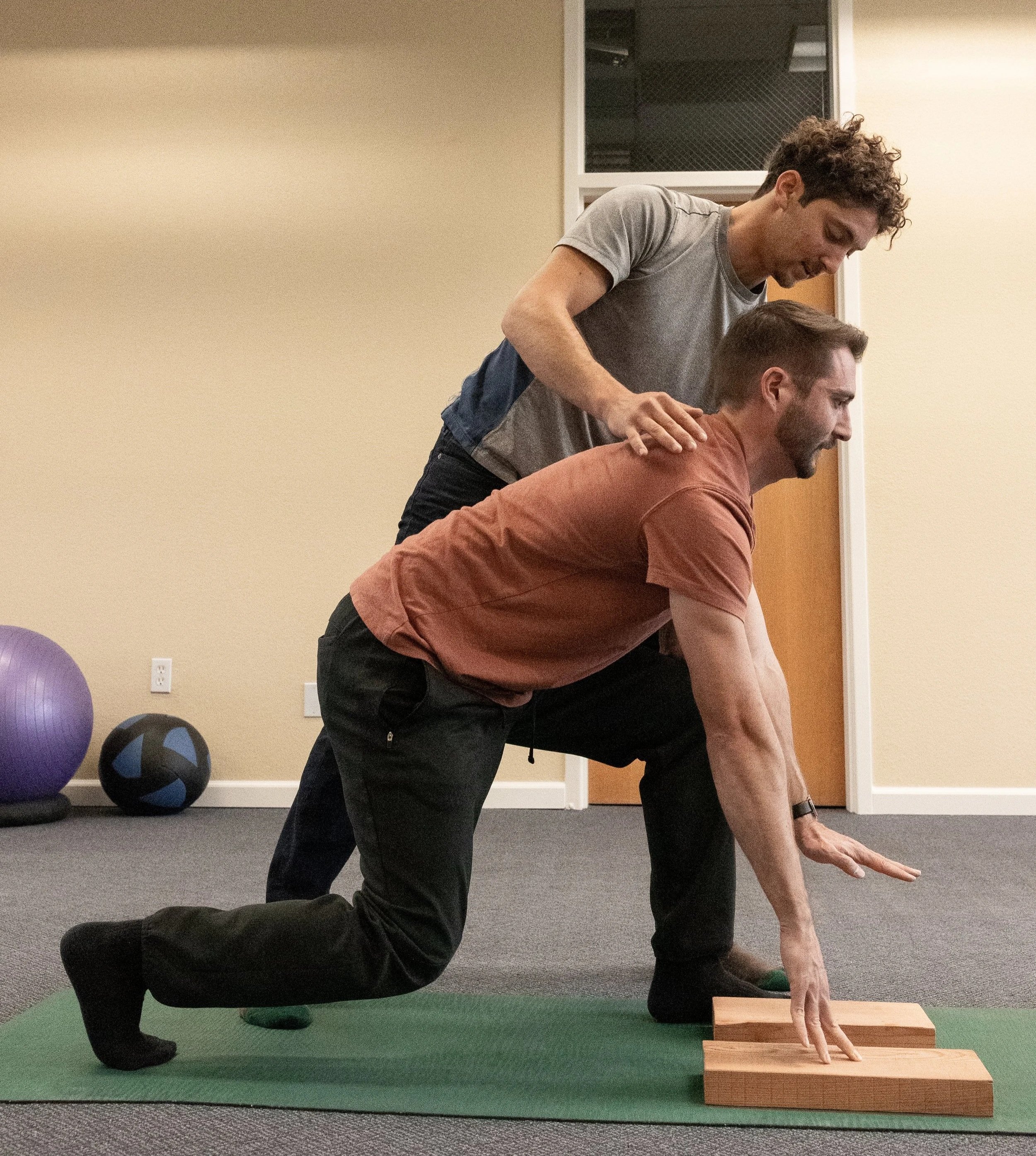 A person performing a bodyweight movement with another person providing support. The person in front is on all fours with their hand on a block, and the person behind is kneeling and leaning forward, hands on the first person's back.