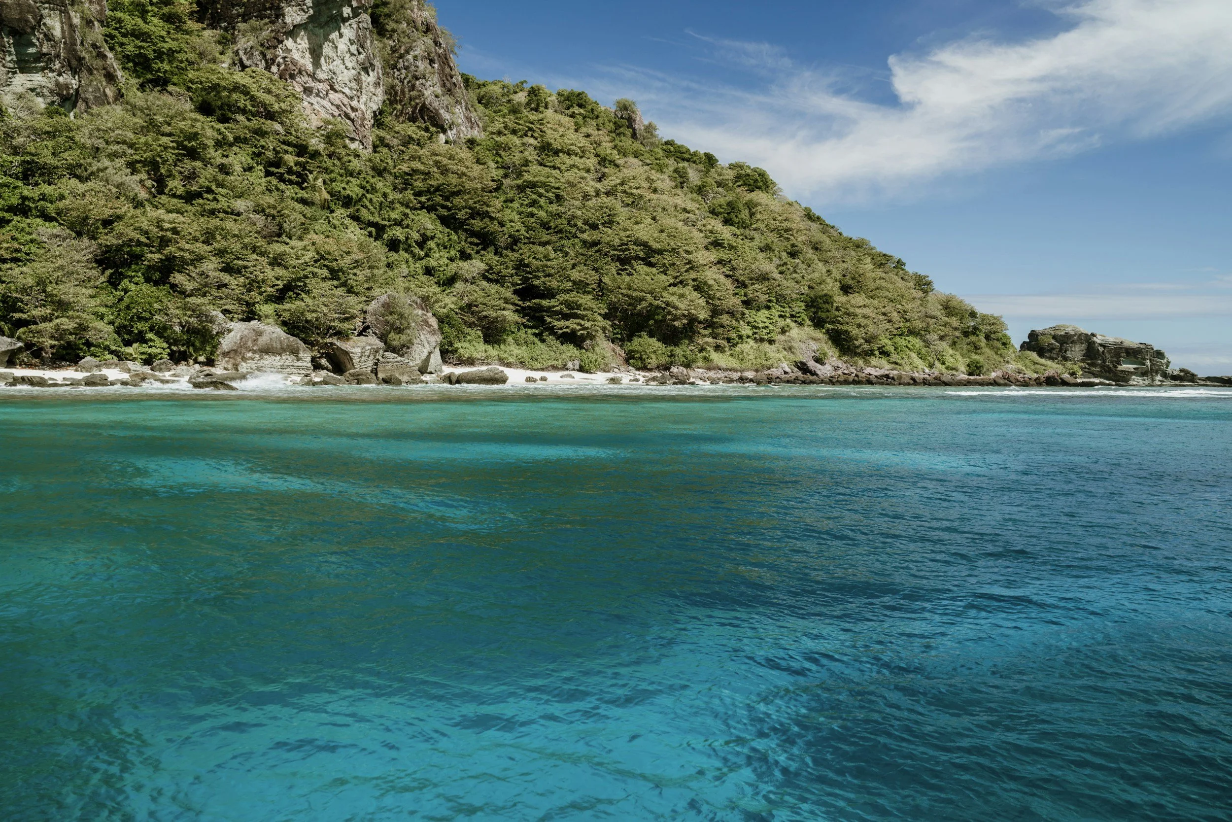 A scenic view of a lush green forested hillside meeting the turquoise water of the ocean, with a partly cloudy sky overhead.