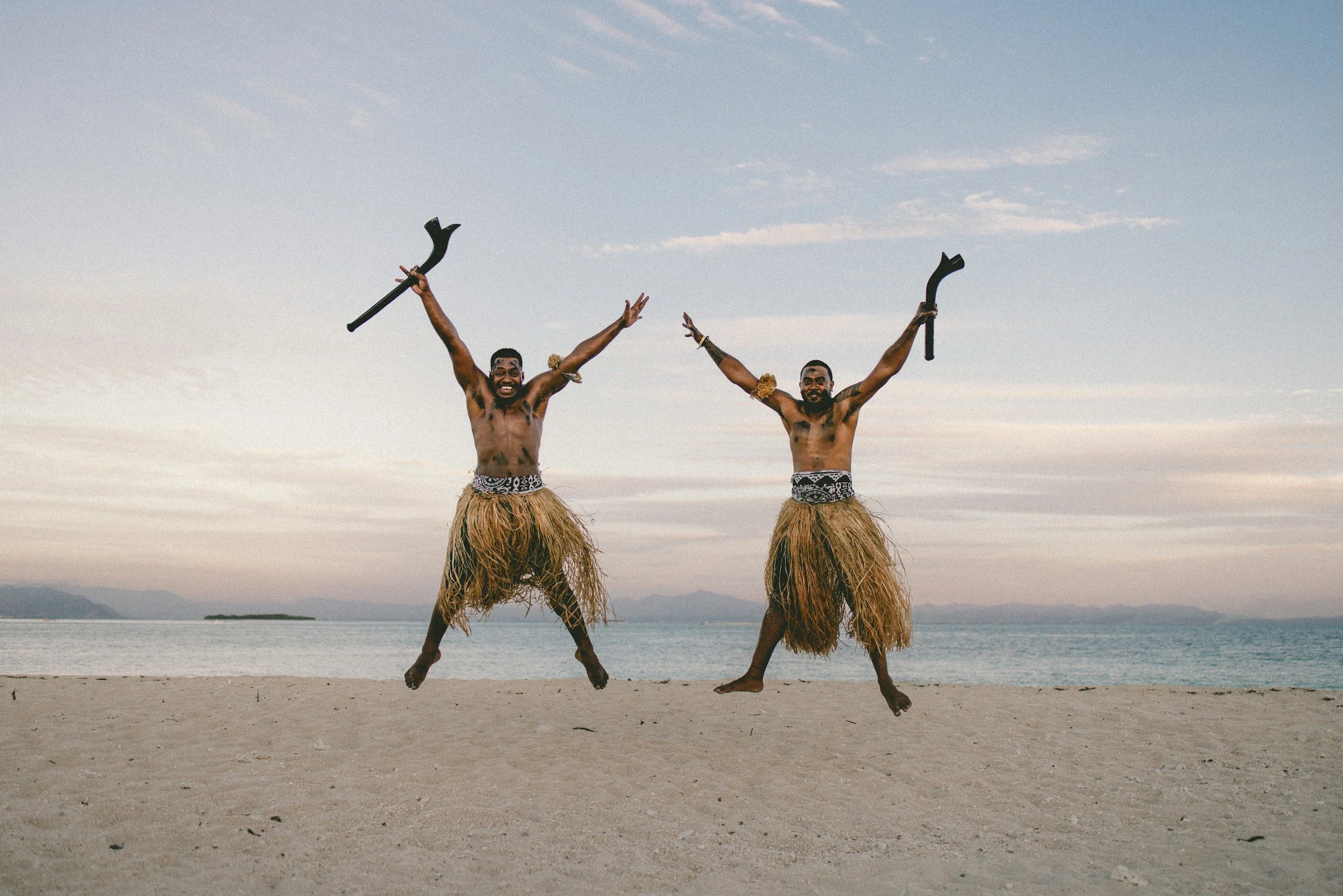 Two men jumping on a beach wearing traditional grass skirts and holding sticks, with arms raised and smiling, against a backdrop of a cloudy sky and water.