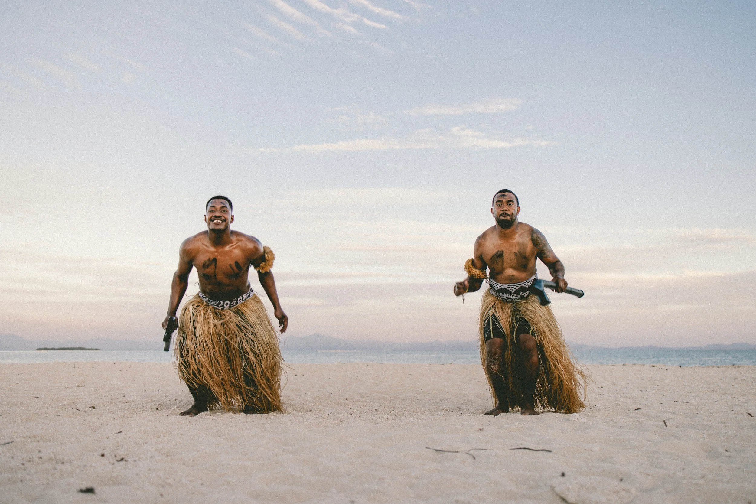Two men dressed in traditional grass skirts and decorated with tribal markings and accessories, standing and smiling on a sandy beach with water and mountains in the background.