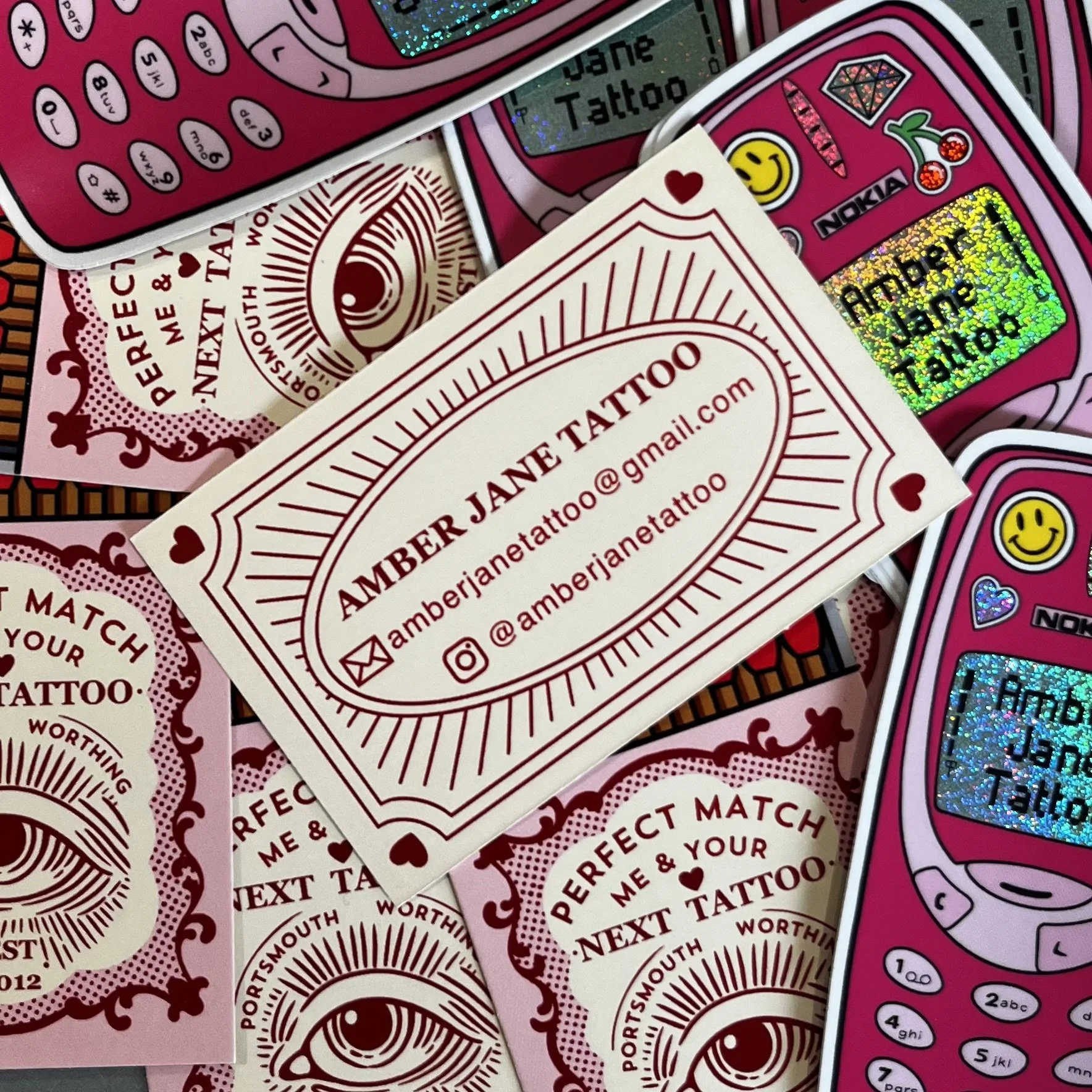 Business card for Amber Jane Tattoo with contact information, surrounded by various colorful stickers and phone-shaped phone cases with stickers.