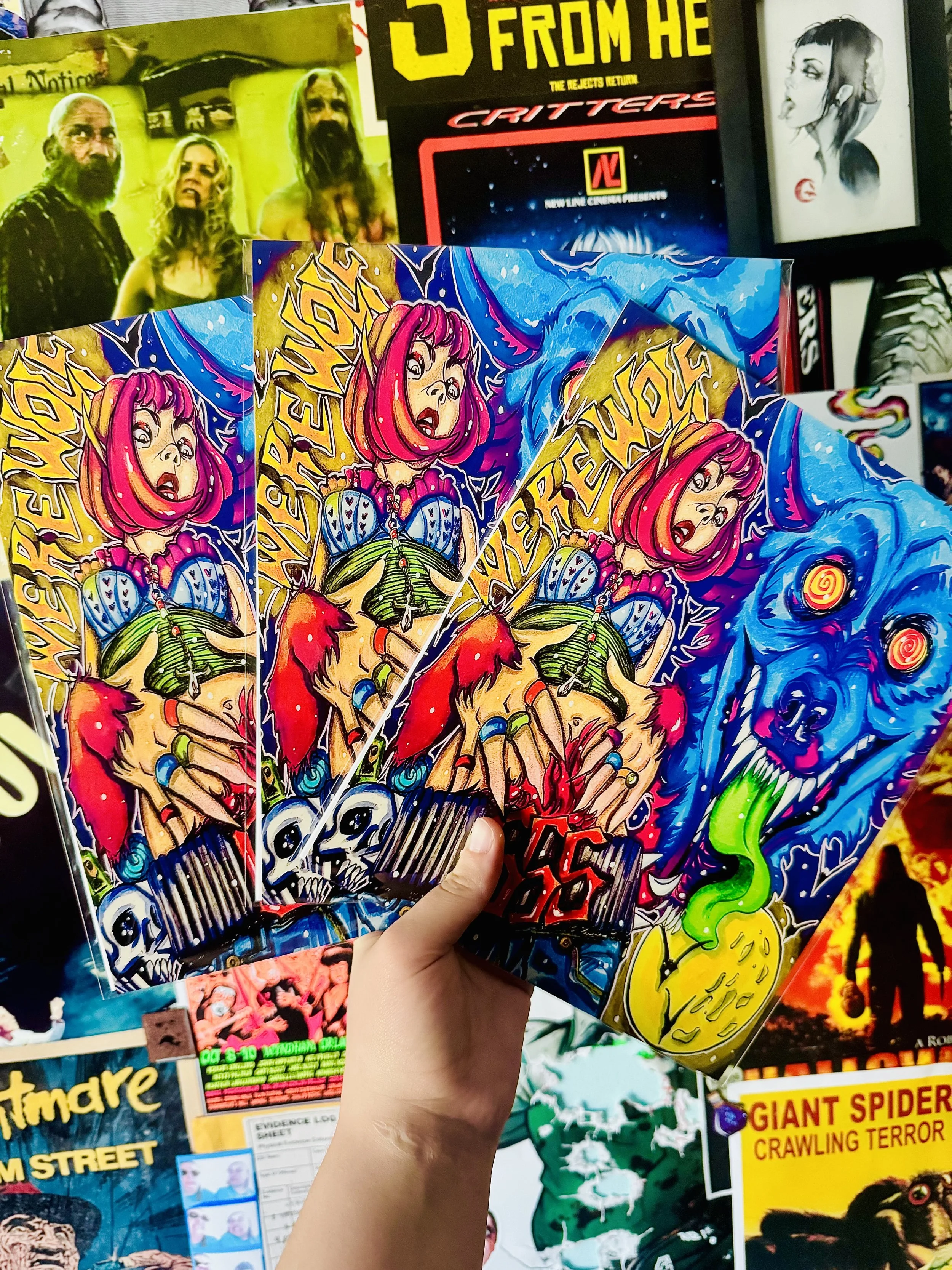 Hand holding multiple colorful comic book covers featuring a surprised girl with pink hair and fantasy elements, with various posters in the background including a black and white portrait and a poster with people making shocked expressions.