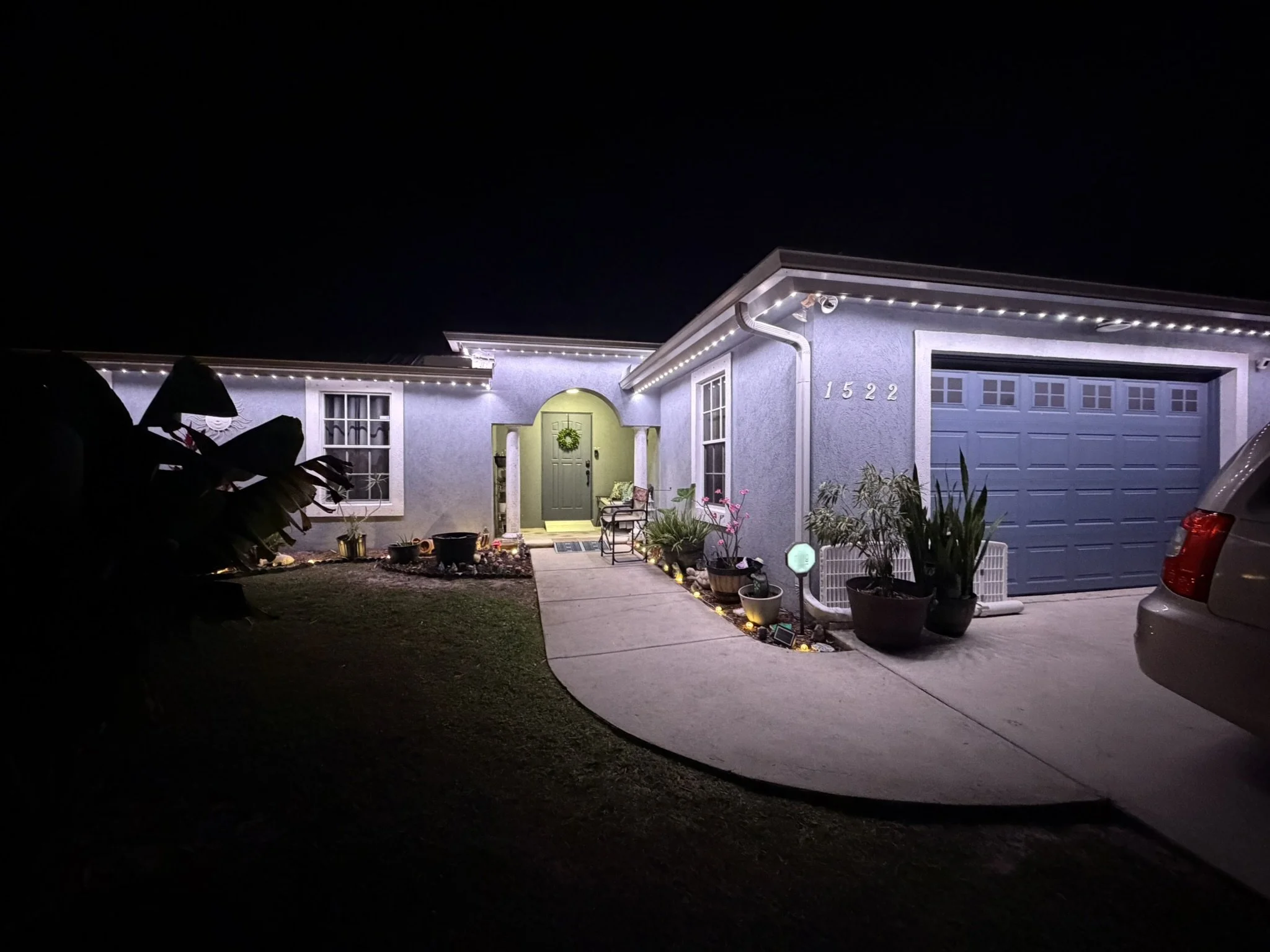 Night view of a house decorated with string lights outlining the roof and around the entrance, with potted plants and outdoor furniture near the front door, and a car parked in the driveway.