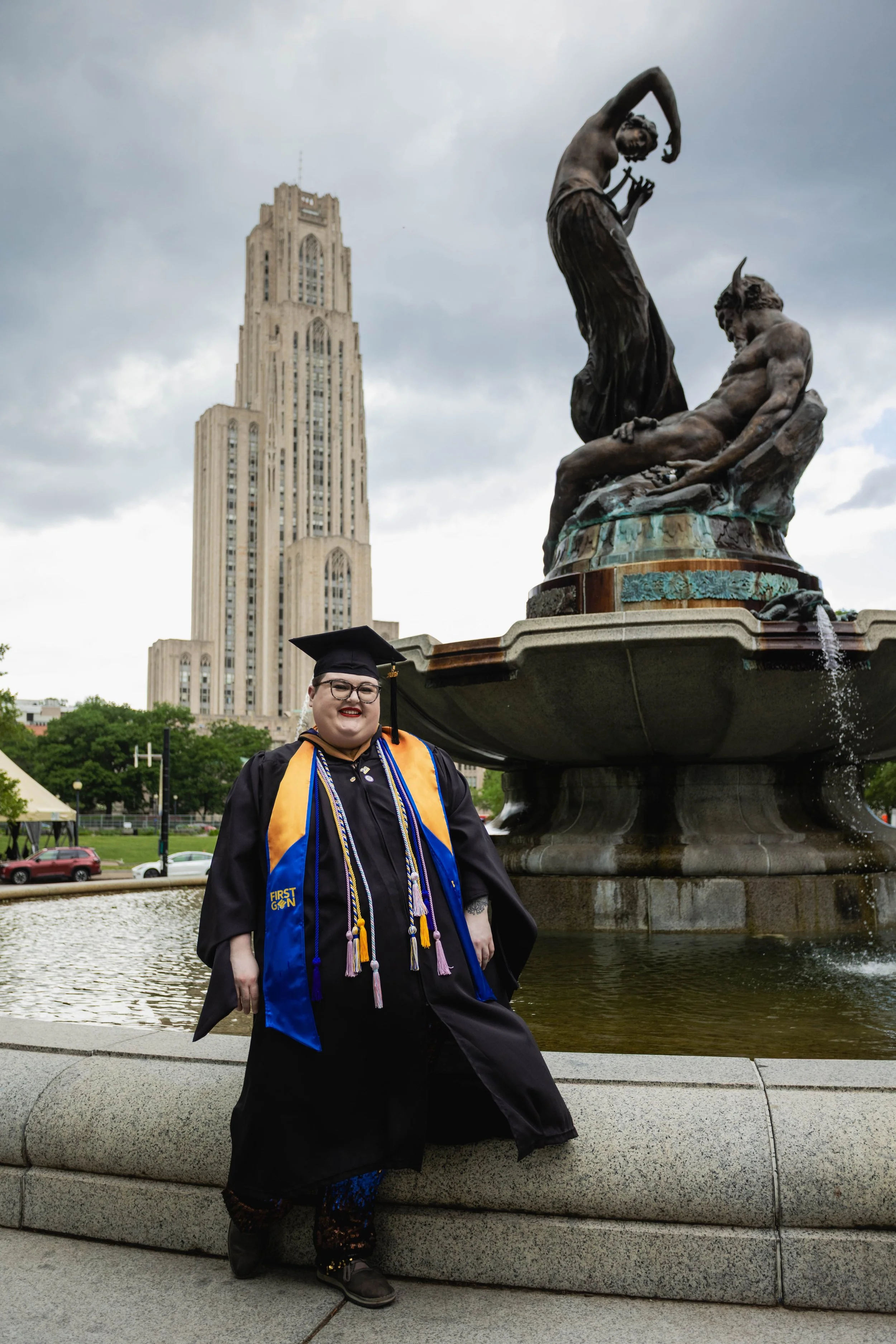 A plus size nonbinary person wearing masters degree graduation regalia stands in front of a fountain and the Cathedral of Learning on the University of Pittsburgh campus in Pittsburgh, Pennsylvania.