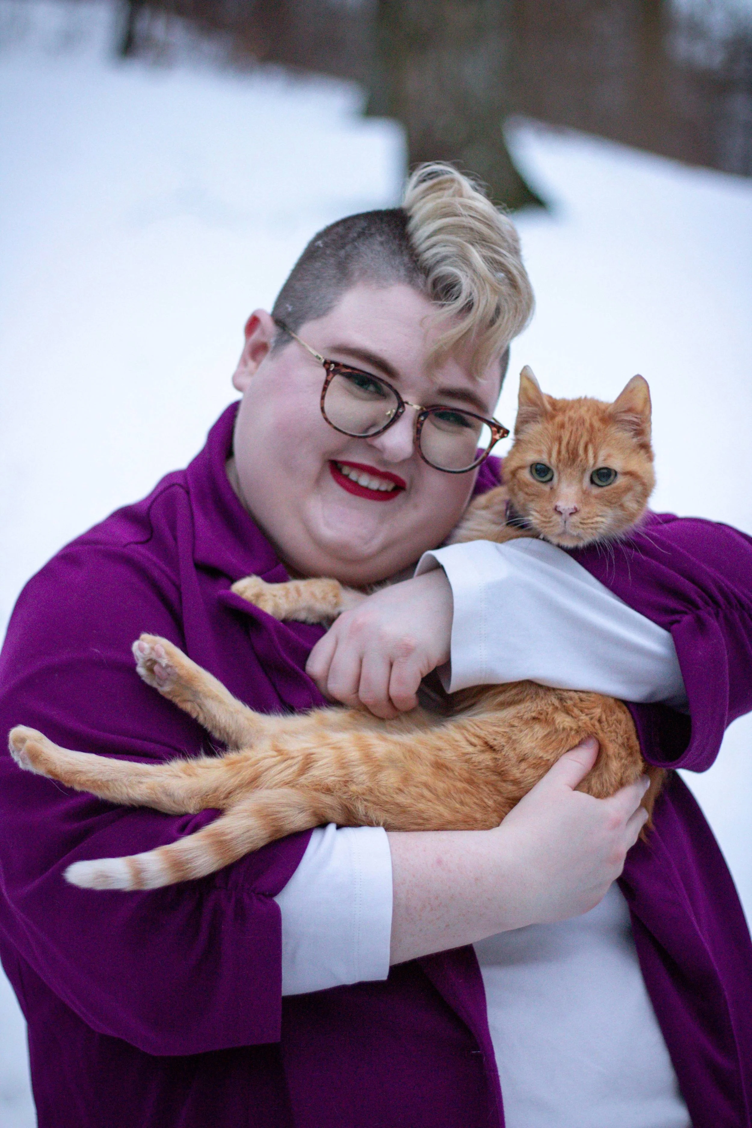A plus-size nonbinary human with shaved hair on the sides and blonde hair on top holds an elderly orange tabby cat. The human is wearing glasses and makeup along with a purple blazer and white shirt against a snow backdrop.