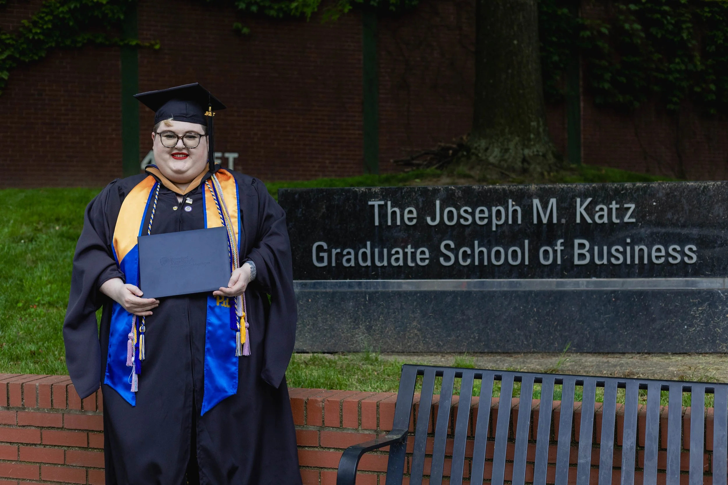 A plus size nonbinary person wearing masters degree graduation regalia holds their degree in front of a sign saying "Joseph M Katz Graduate School of Business" in Pittsburgh, Pennsylvania.
