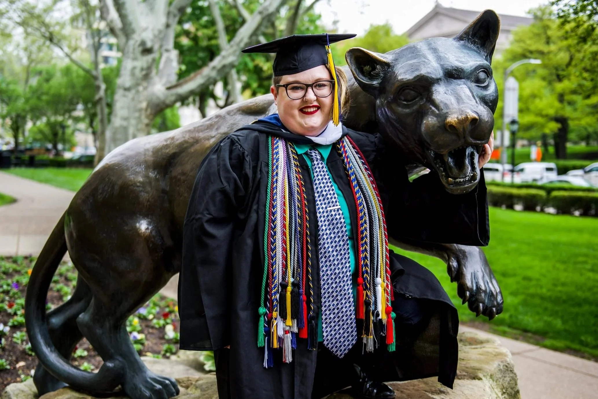 A plus size white person wearing graduation regalia and honor cords from the University of Pittsburgh stands in front of a panther statue, smiling.