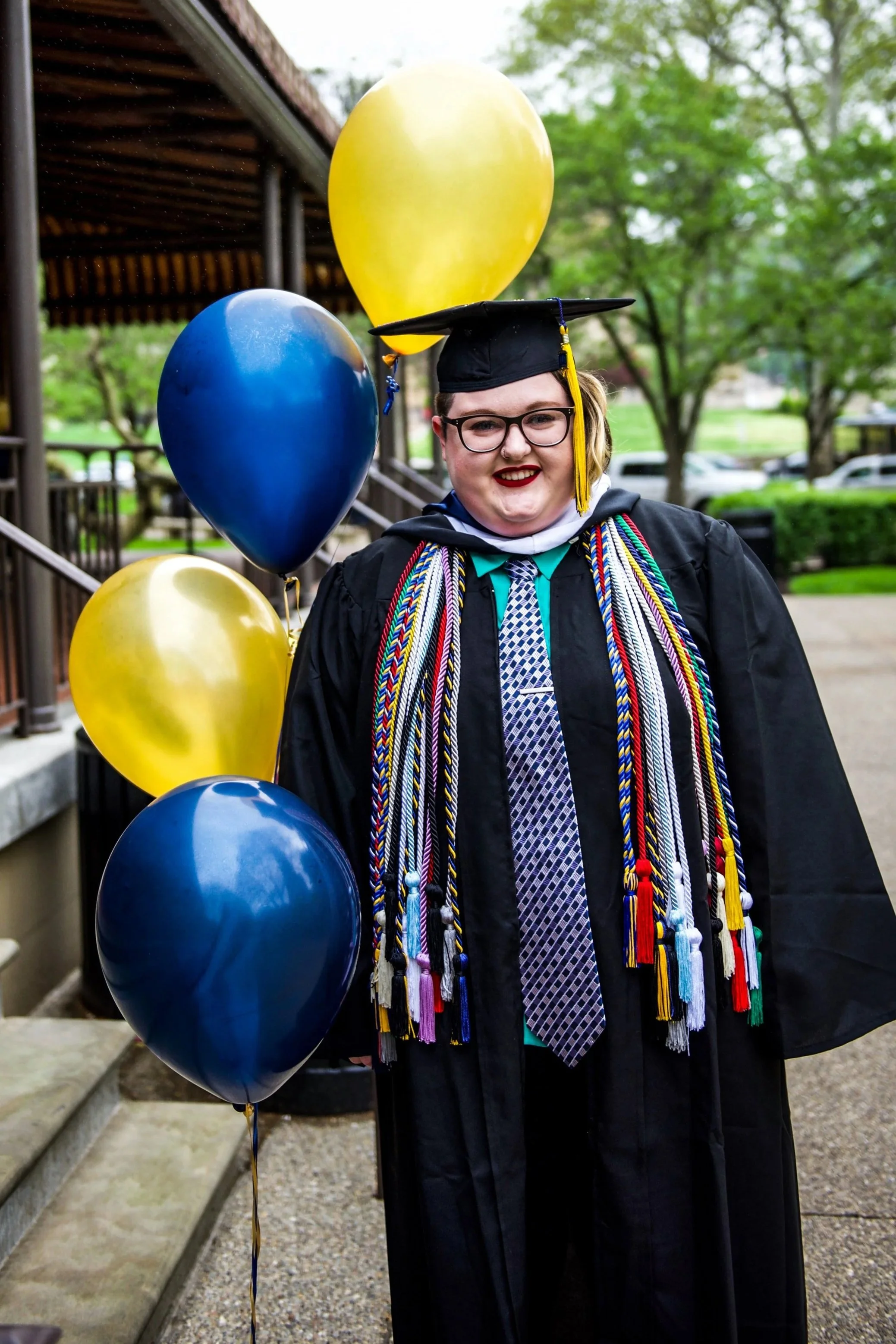 A person stands in front of blue and gold balloons proudly wearing university of Pittsburgh graduation regalia. They are wearing a cap, gown, and many honor cords.