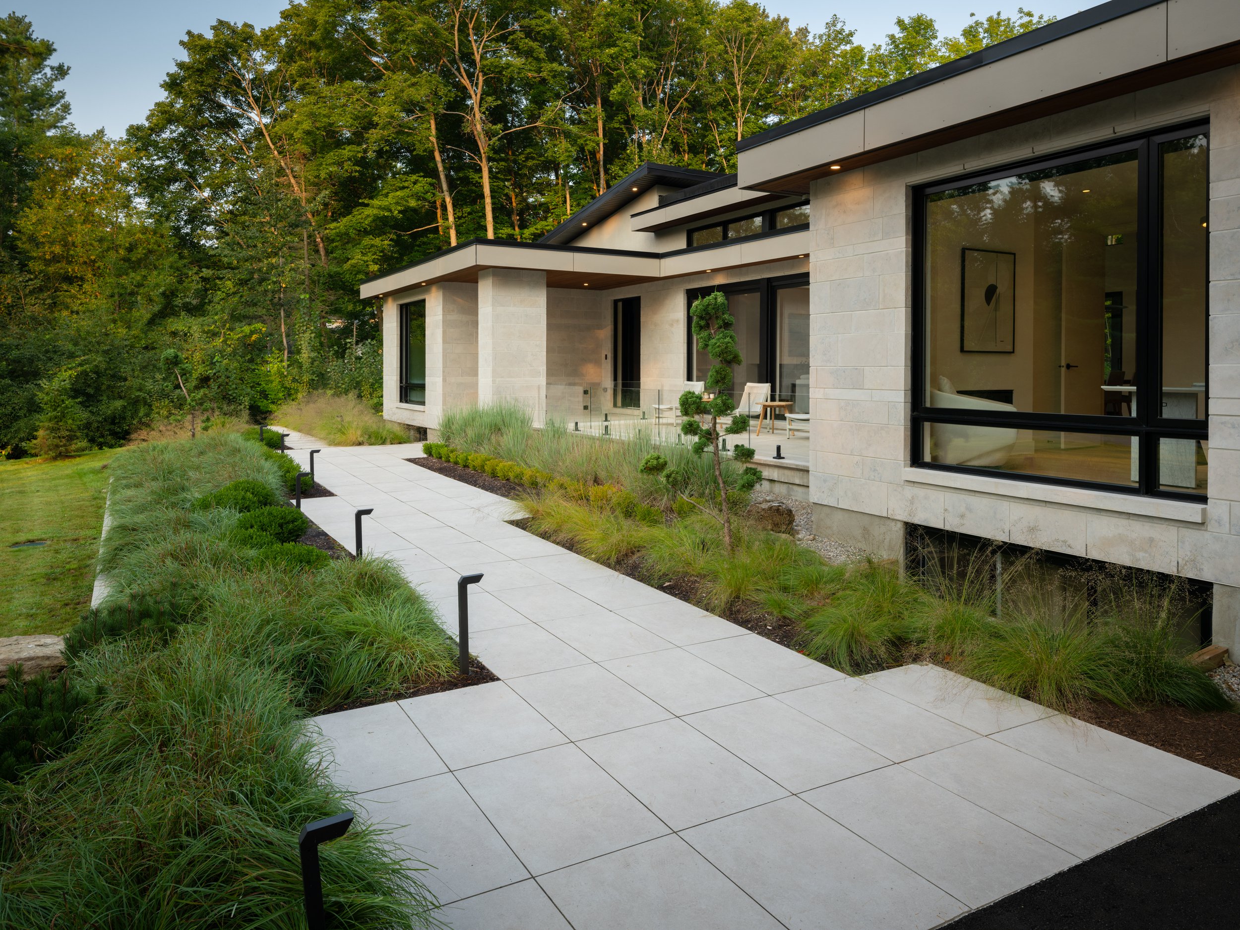 Modern house with large windows, surrounded by greenery. A tiled walkway runs through the landscaped garden, leading to the entrance.