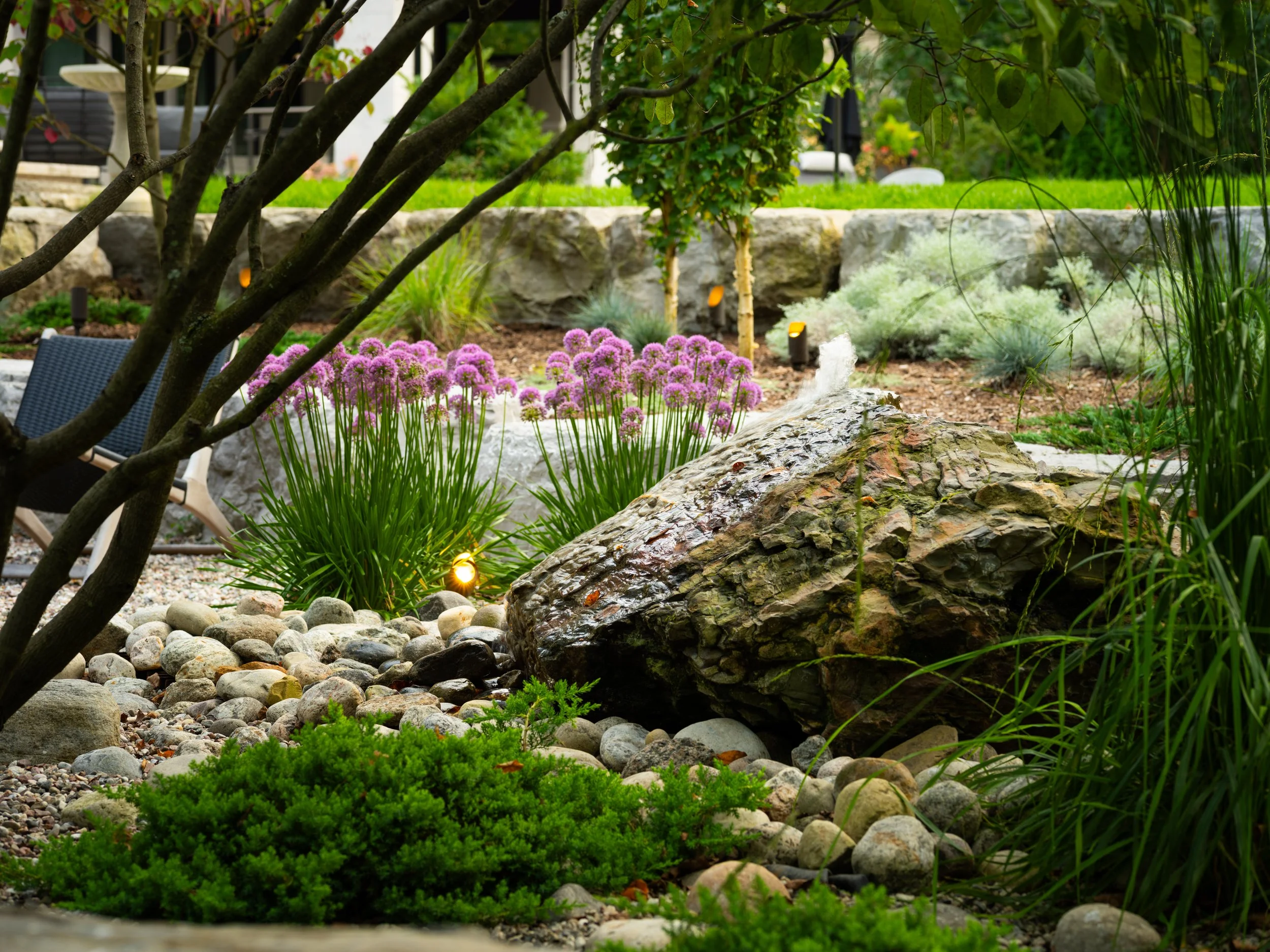 A landscaped garden crafted by a skilled landscape designer features a large rock, purple flowers, shrubs, and small stones. Trees and plants provide a lush backdrop, with pathway lights visible throughout.