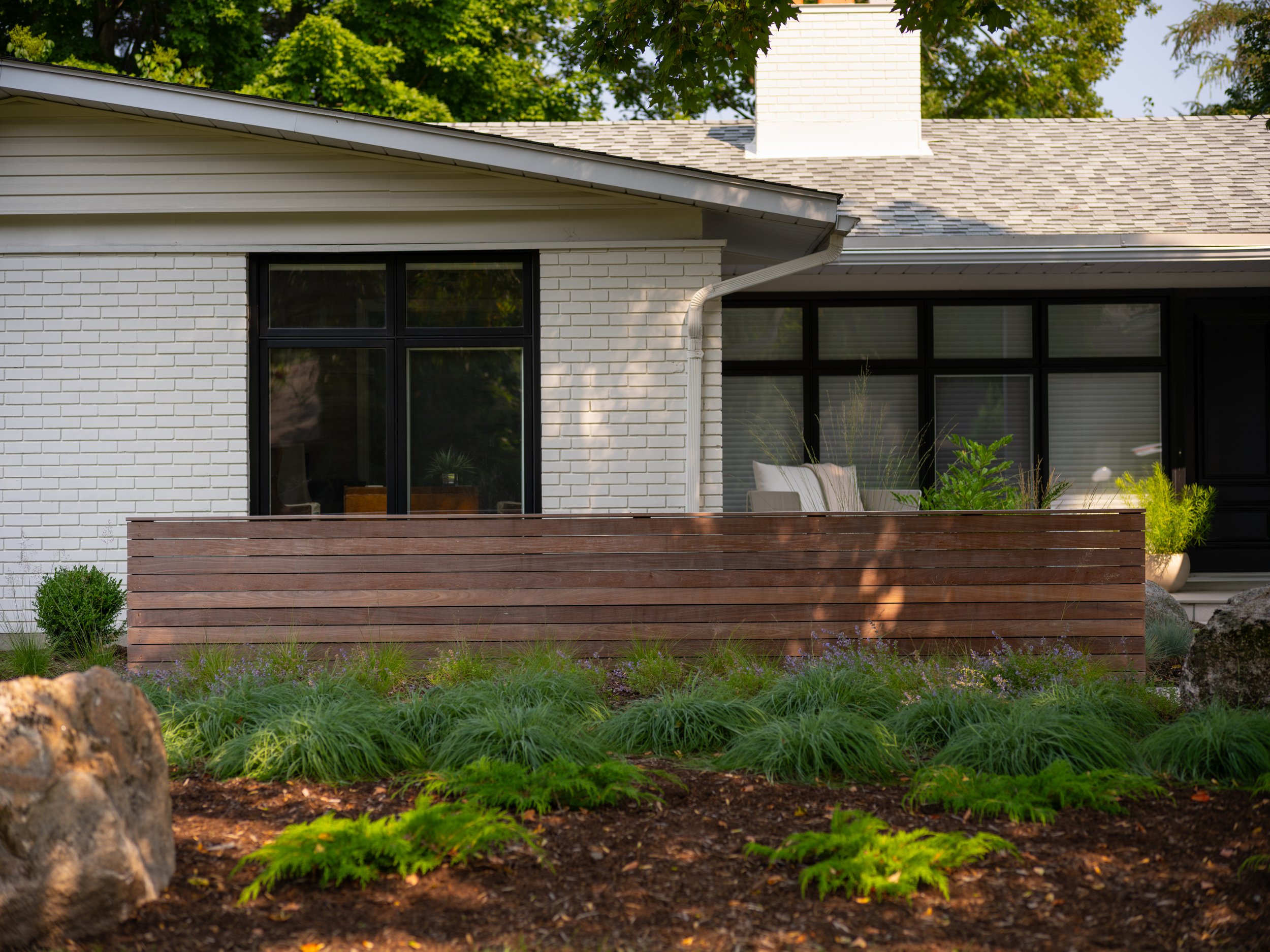 A modern house with white brick walls and large windows is harmoniously integrated into the landscape design, featuring a wooden fence surrounded by lush green shrubs and plants under a clear sky.