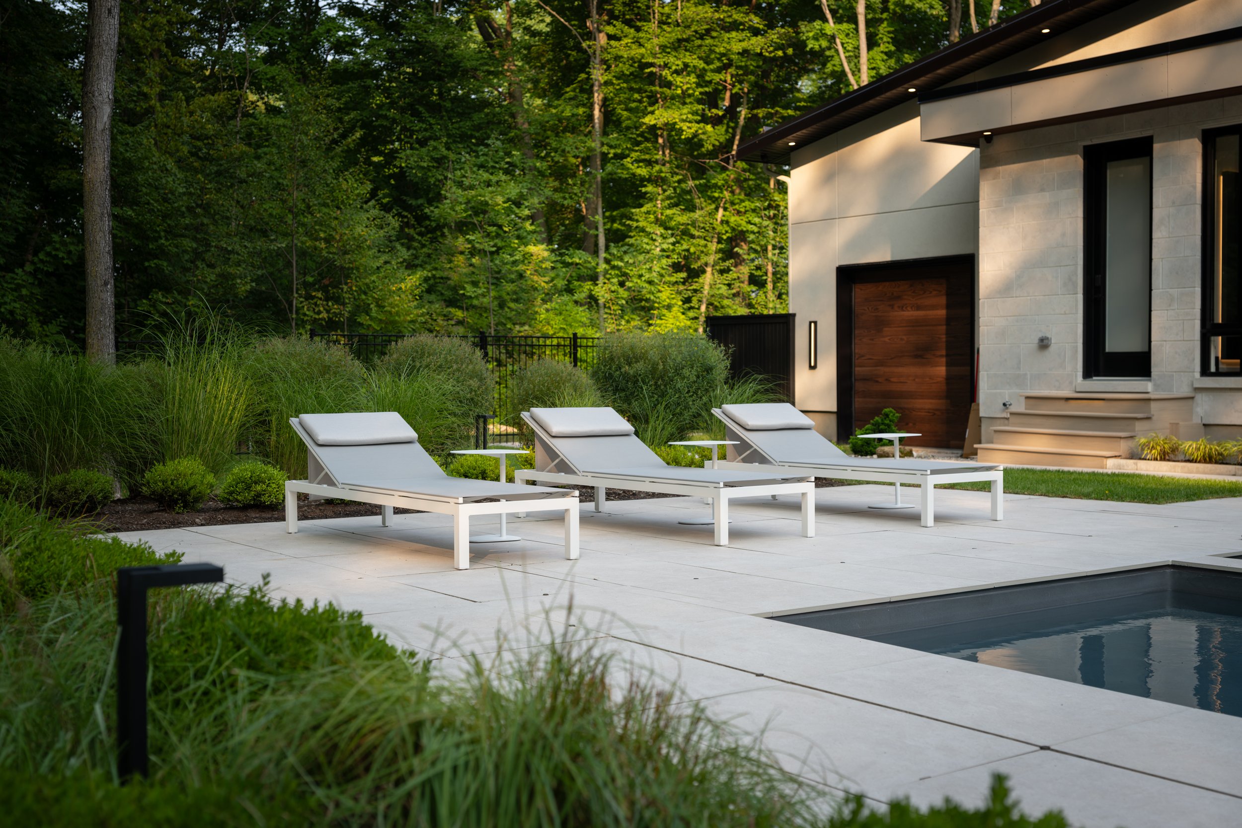 Three white loungers rest beside a modern house's pool, seamlessly integrated into an exquisite landscape design, surrounded by lush greenery and trees.