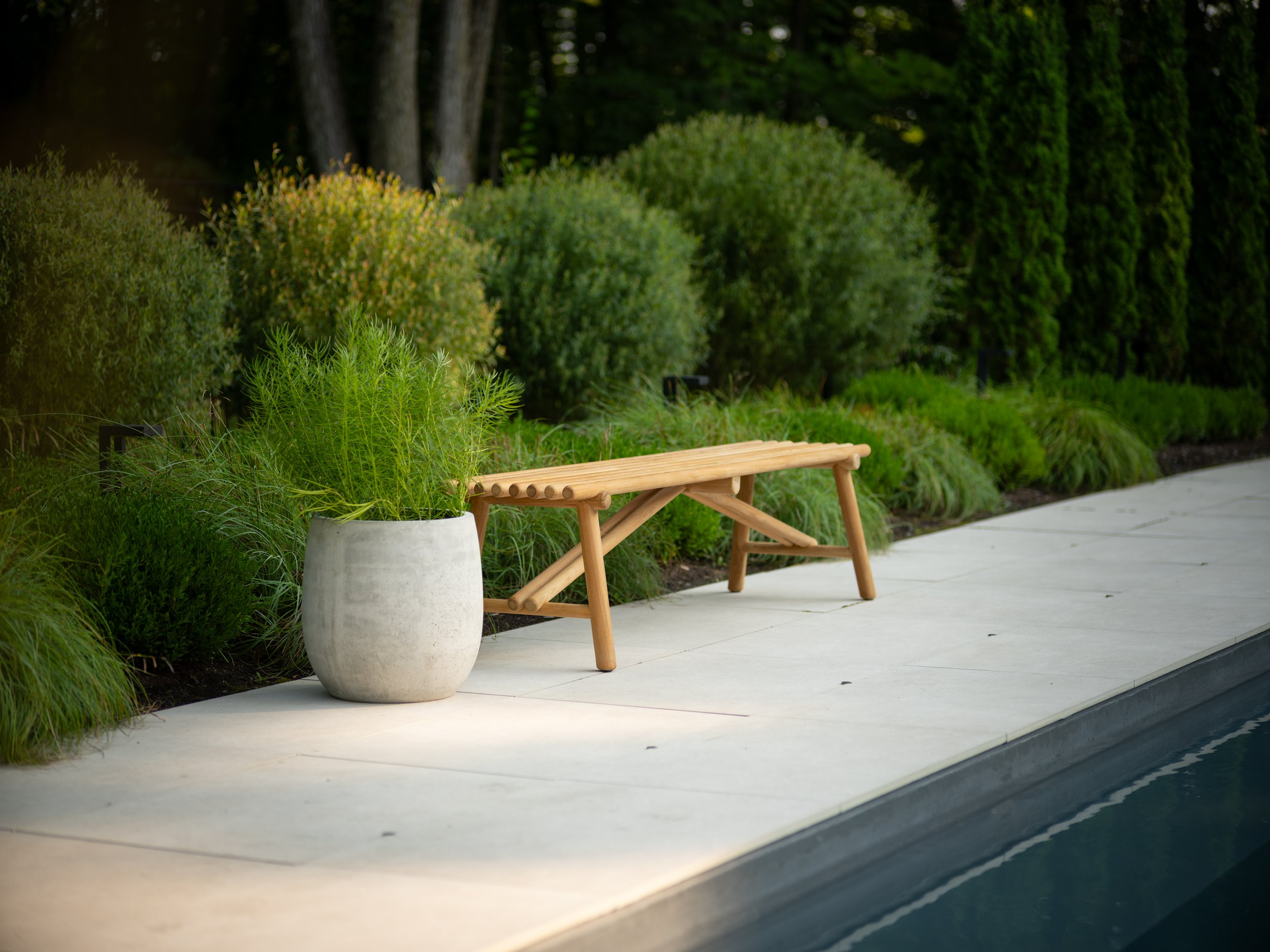 A beautifully crafted wooden bench and large potted plant enhance the poolside landscape design, surrounded by lush greenery and majestic trees.