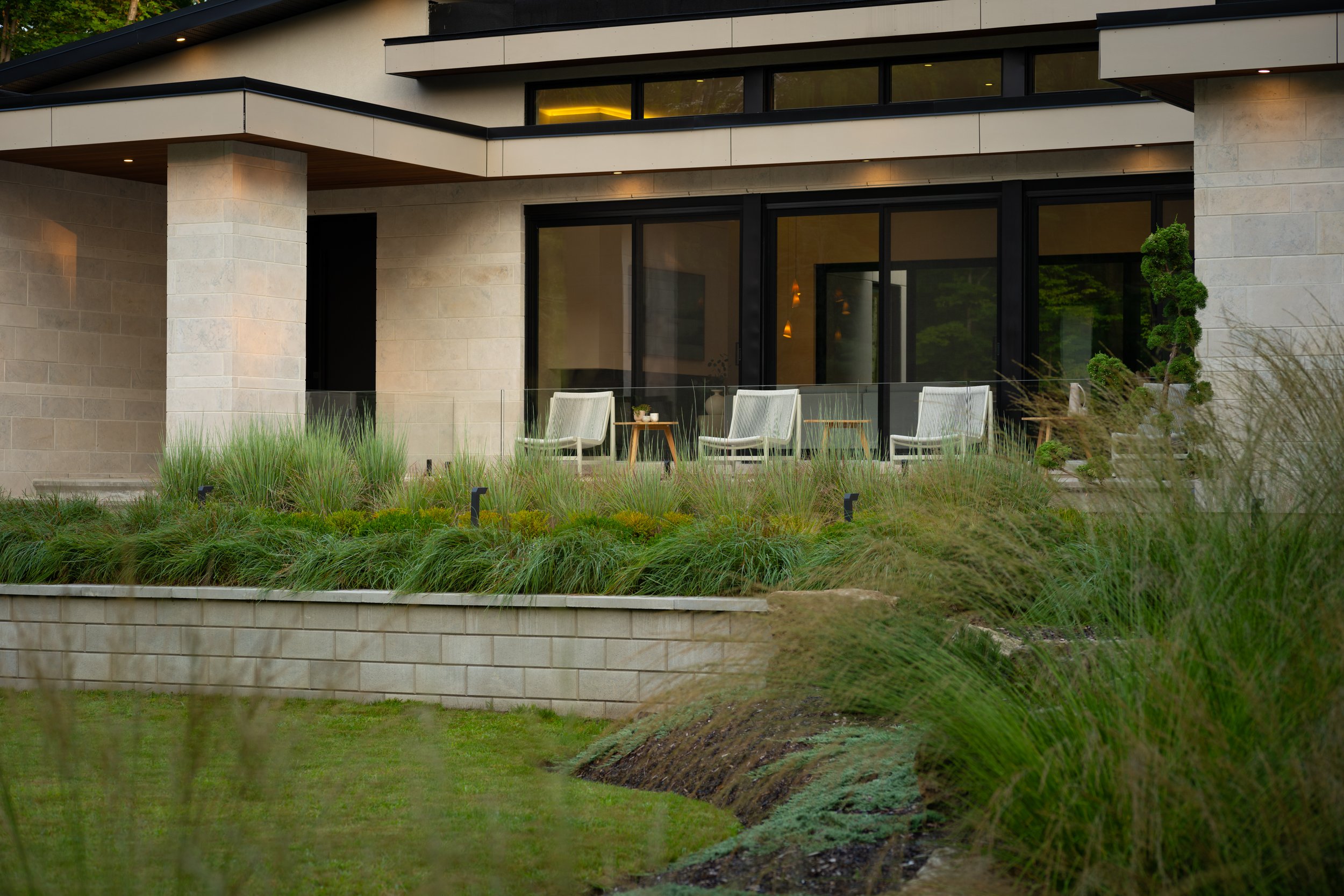 Modern house exterior with large windows and a patio featuring two white chairs. The foreground has landscaped greenery and grass.