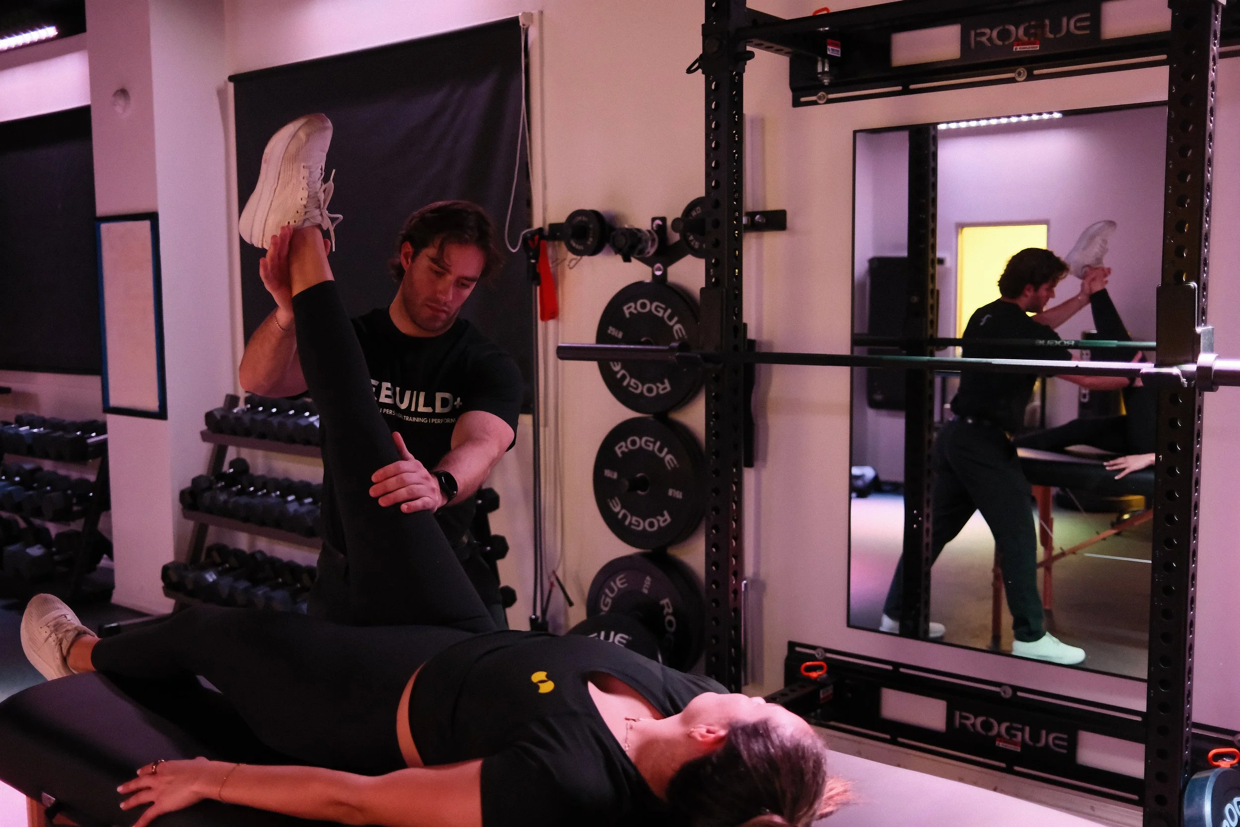 Physical Therapist assisting a woman with a stretching exercise in a gym, with gym equipment and a mirror in the background.