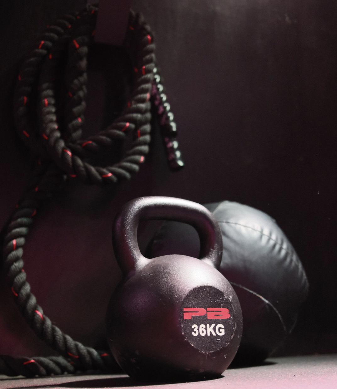 Black kettlebell labeled 36 kilograms, coiled battle ropes with red accents, and a medicine ball against a dark wall.