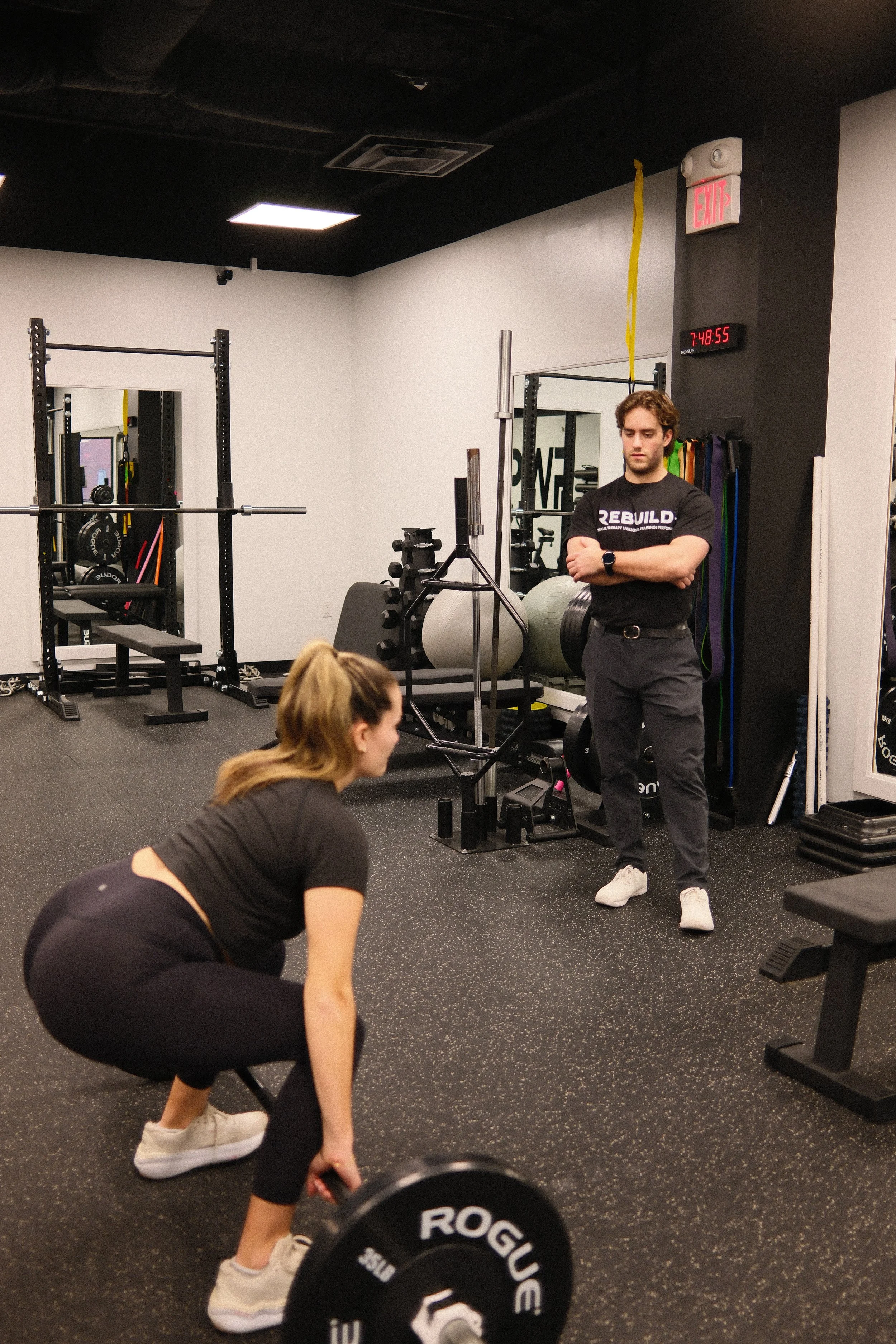 A woman is lifting a barbell in a gym while a personal trainer observes. The gym has weightlifting equipment and motivational signage.