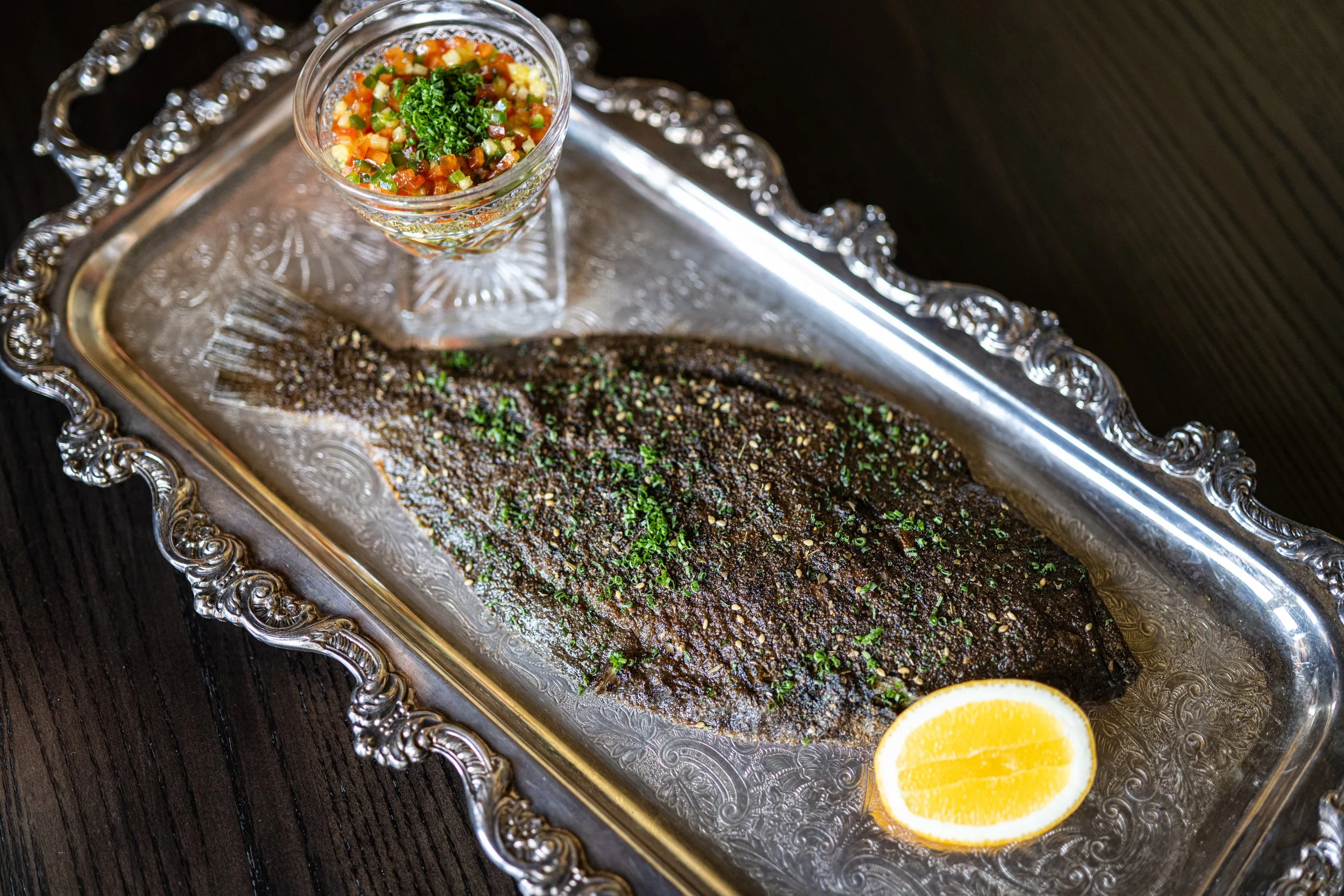 A whole grilled fish topped with herbs on an ornate silver platter, garnished with a lemon wedge, served alongside a small side dish
