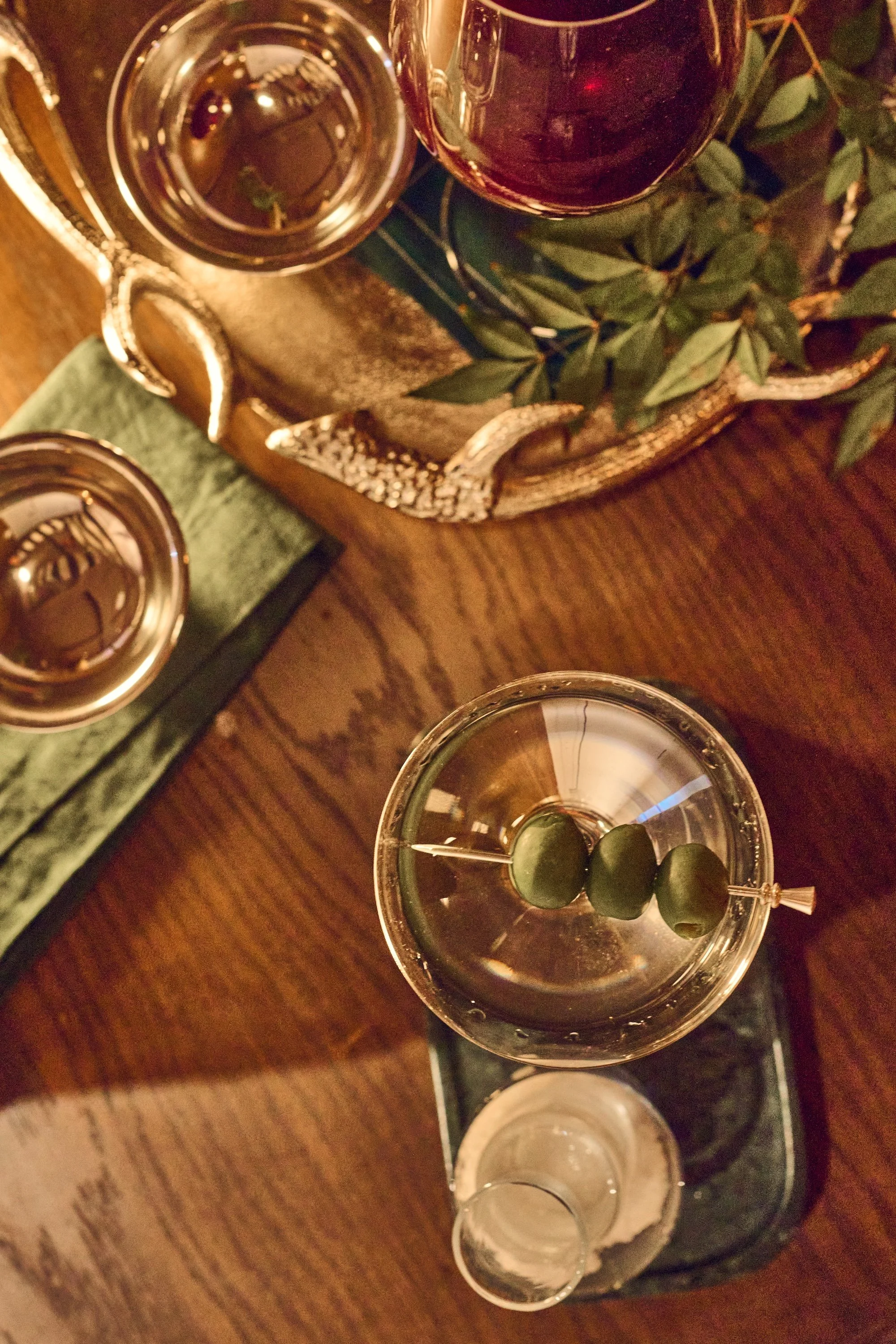 Top-down view of three glasses of wine, one with green olives, on a wooden table with a tray of greenery and a decorative tray.