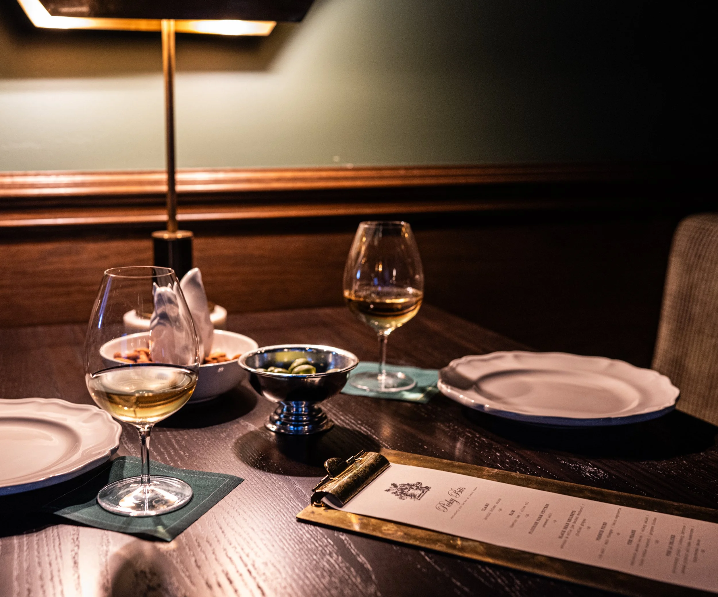 A dimly lit dining table with two wine glasses filled with white wine, a small bowl of olives, a decorative menu on a clip, and some plates with napkins inside a cozy restaurant setting.