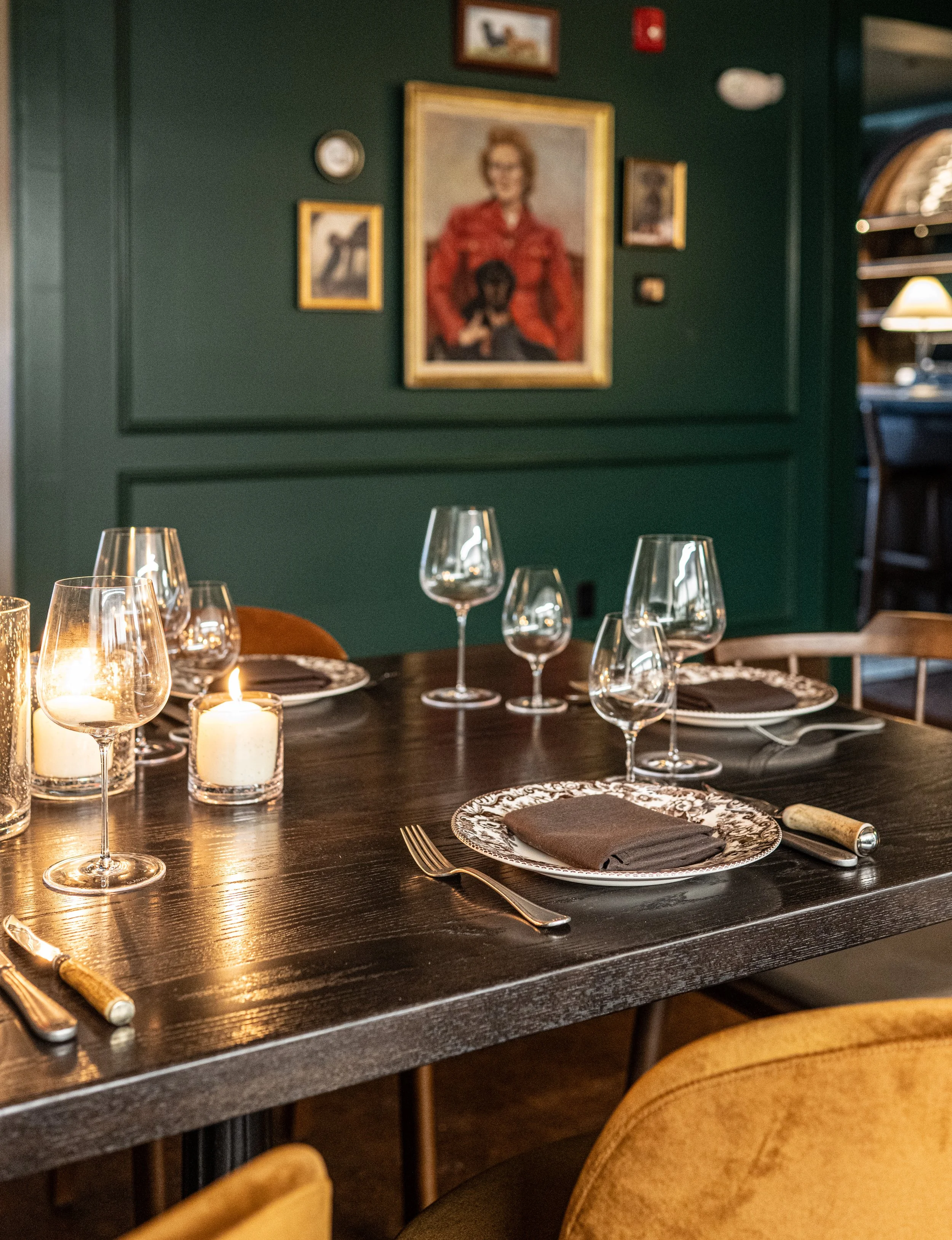 A dining table set for a meal with wine glasses, candles, plates with black napkins, forks, and knives, in a restaurant with dark wood furniture and art on a green wall.