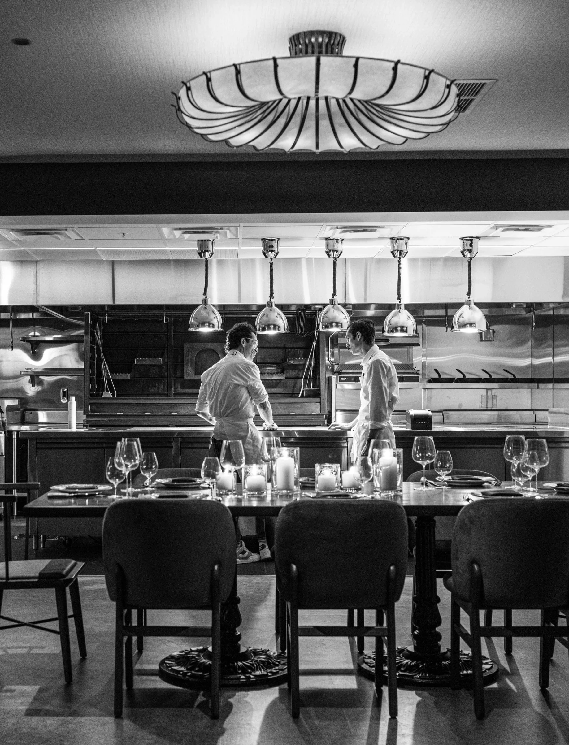 Two chefs in white uniforms working in a professional kitchen, seen through a dining area with a table set with candles and wine glasses.