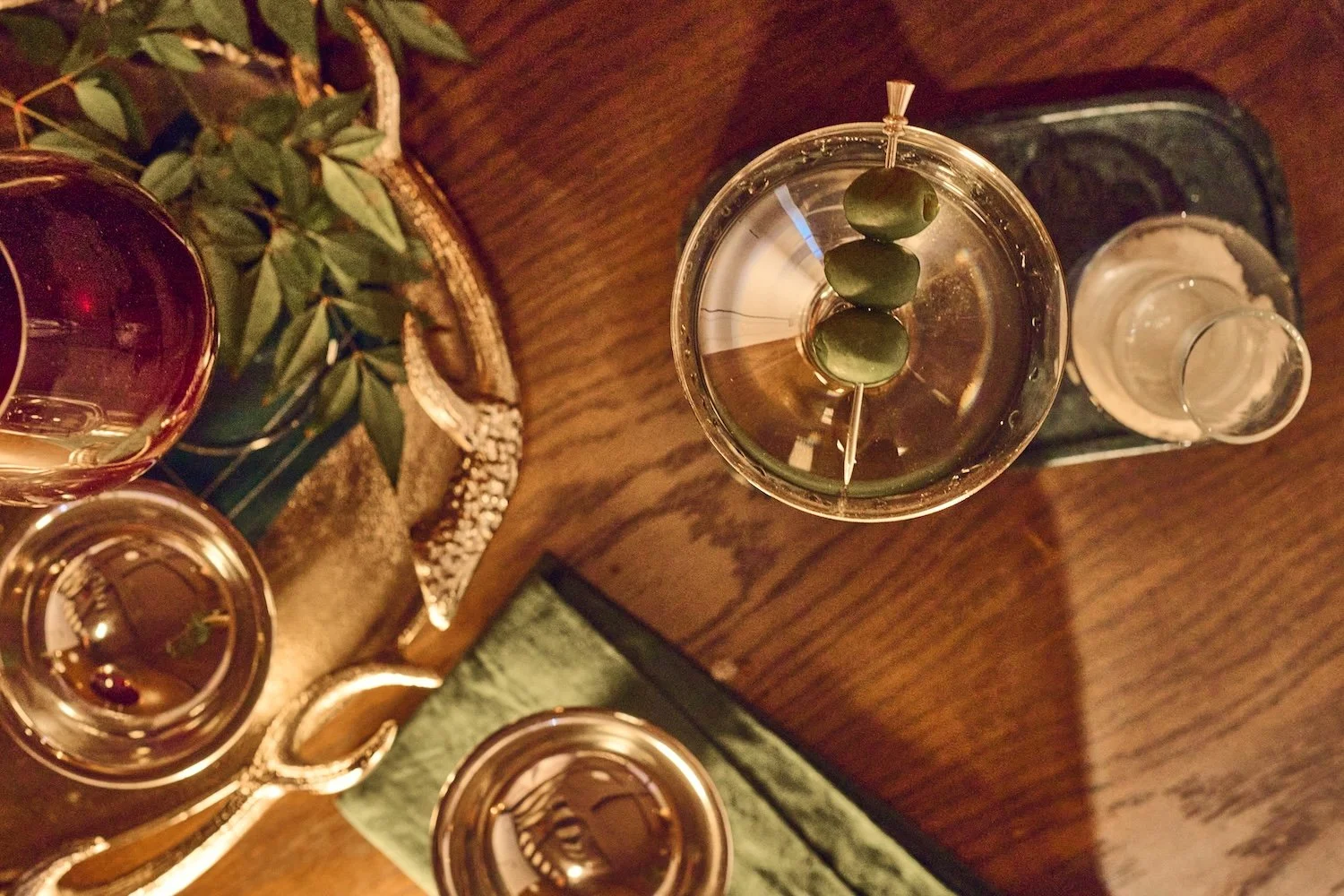 Top-down view of a cocktail glass with three green olives on a skewer and a foggy glass of water behind it, on a wooden table with a green napkin.