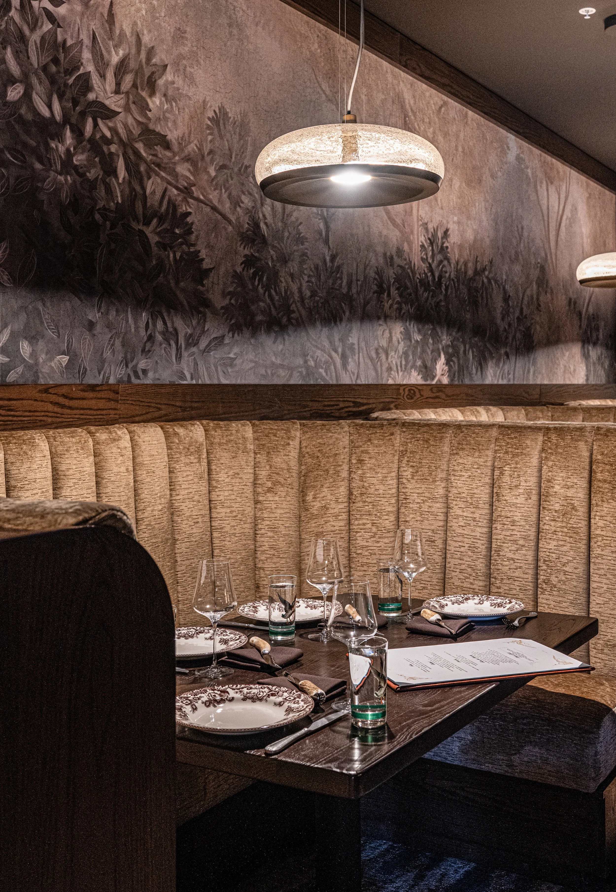 A restaurant booth with a wooden table set for four, decorated with patterned plates, wine glasses, water glasses, and black napkins with utensils. Overhead, modern circular pendant lights hang, and a mural with foliage is on the wall behind the booth.