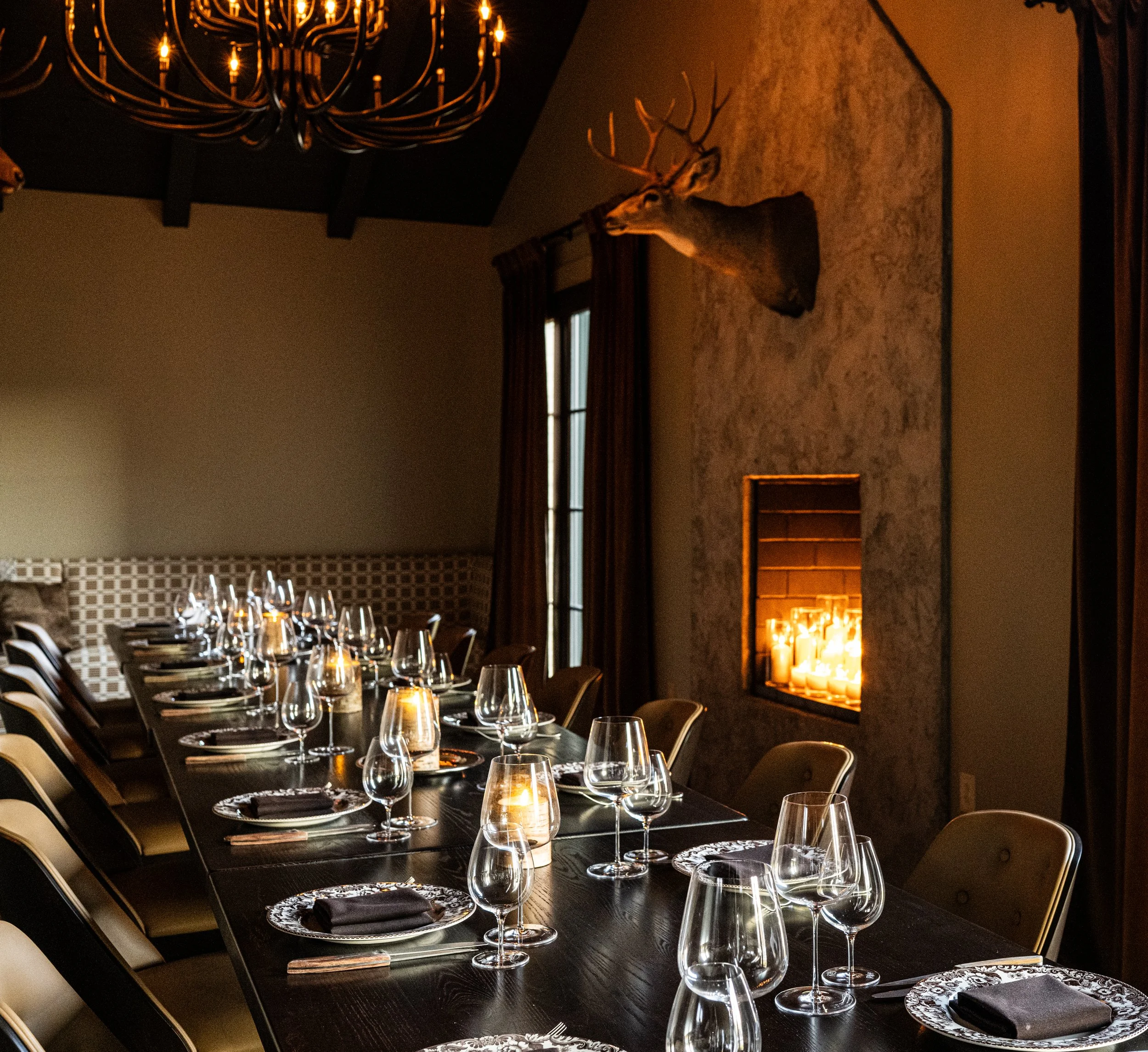 Elegant dining table set for a formal dinner, with wine glasses, black napkins, and decorative plates, in a dimly lit room featuring a fireplace with candles and a mounted deer head on the wall.