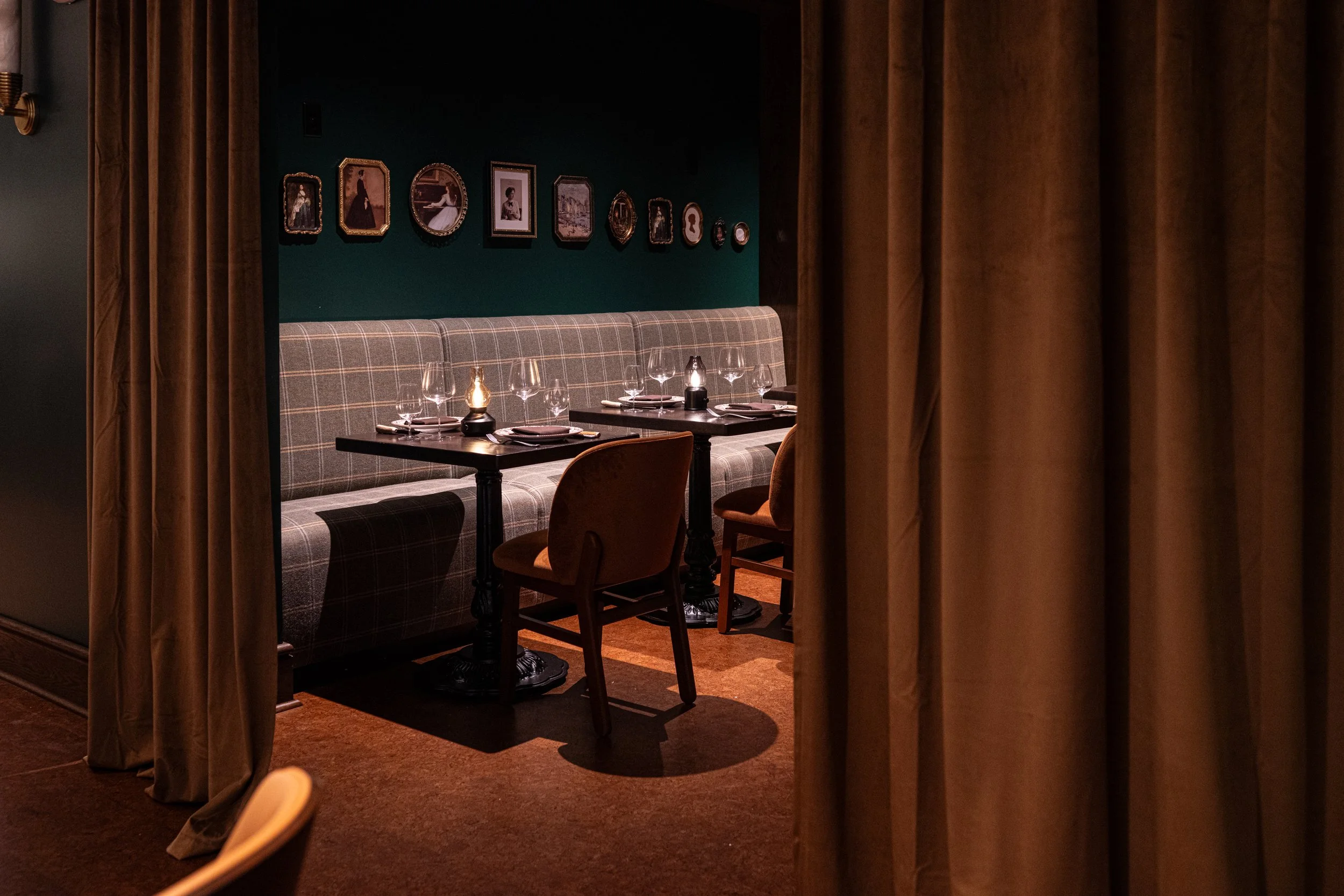 A cozy, dimly lit restaurant booth with a plaid cushioned bench, wooden chairs, tables set with wine glasses, dinner plates, and small lamps, decorated with framed vintage photos on a dark green wall.
