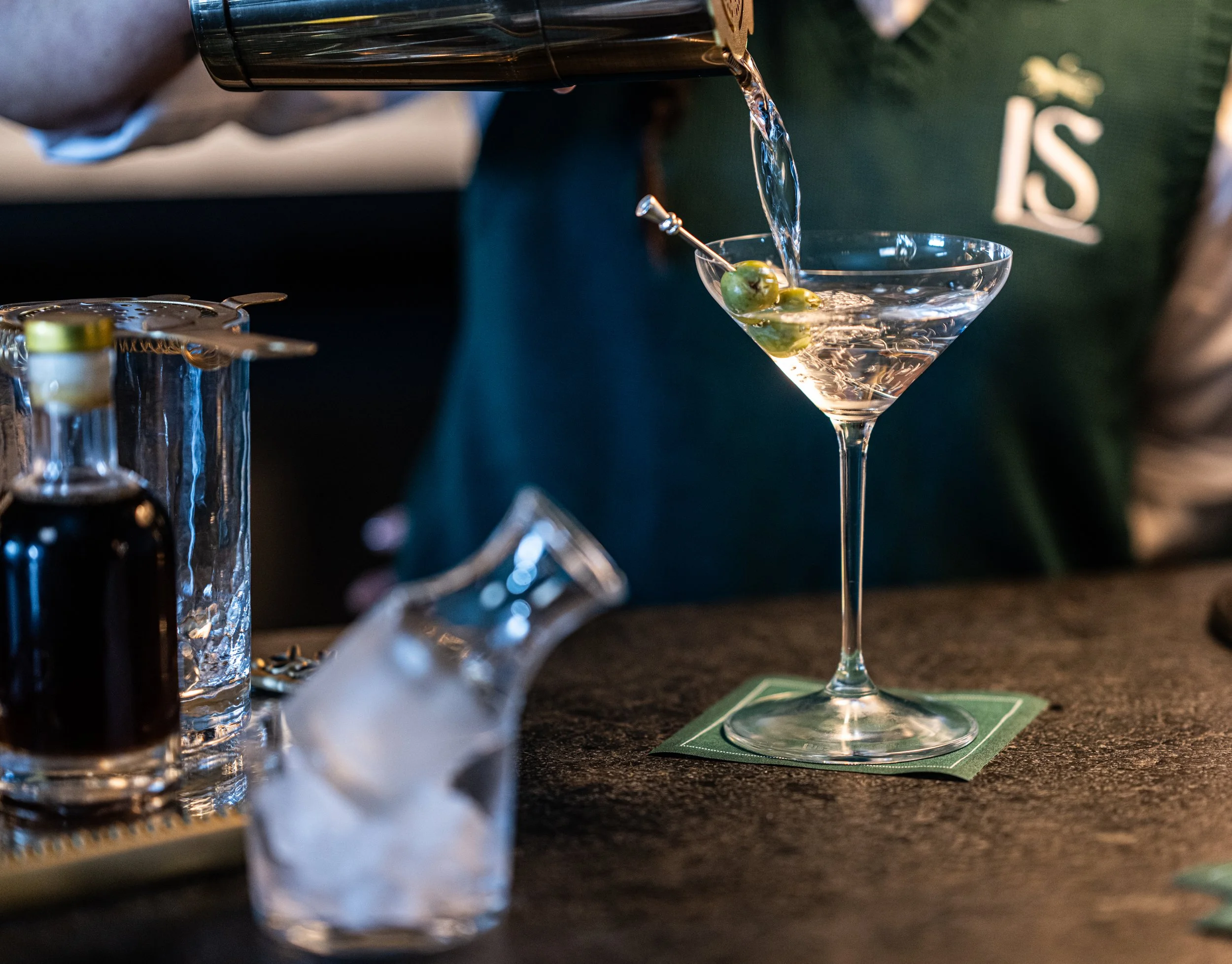 A bartender pouring clear liquid into a martini glass with green olives, on a dark countertop with bar tools and alcohol bottles nearby.