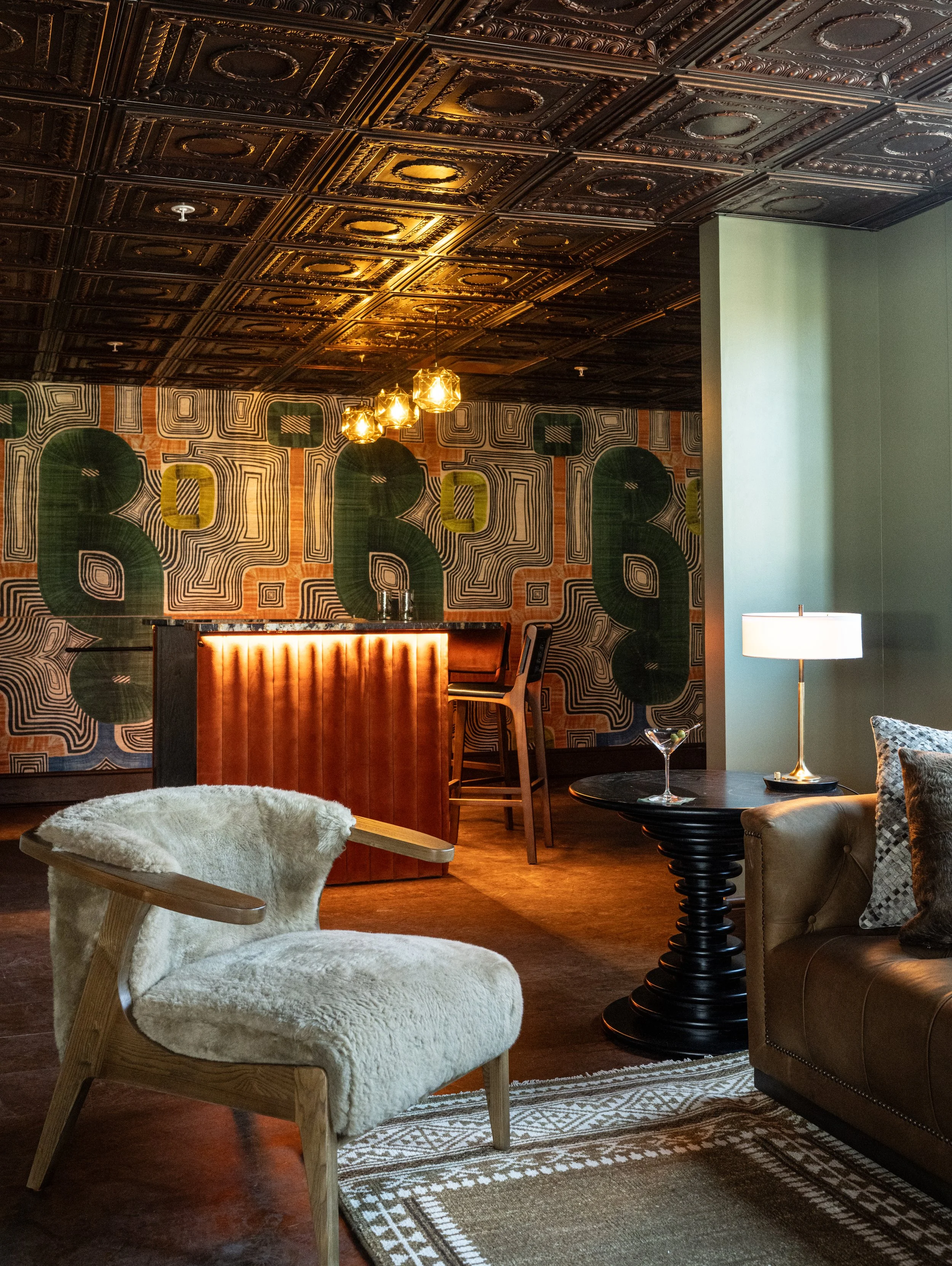 Interior of a stylish lounge with patterned wallpaper, a wooden bar counter, modern lighting fixtures, a plush lounge chair, a black side table with a lamp and a martini glass, and a sofa with decorative pillows.