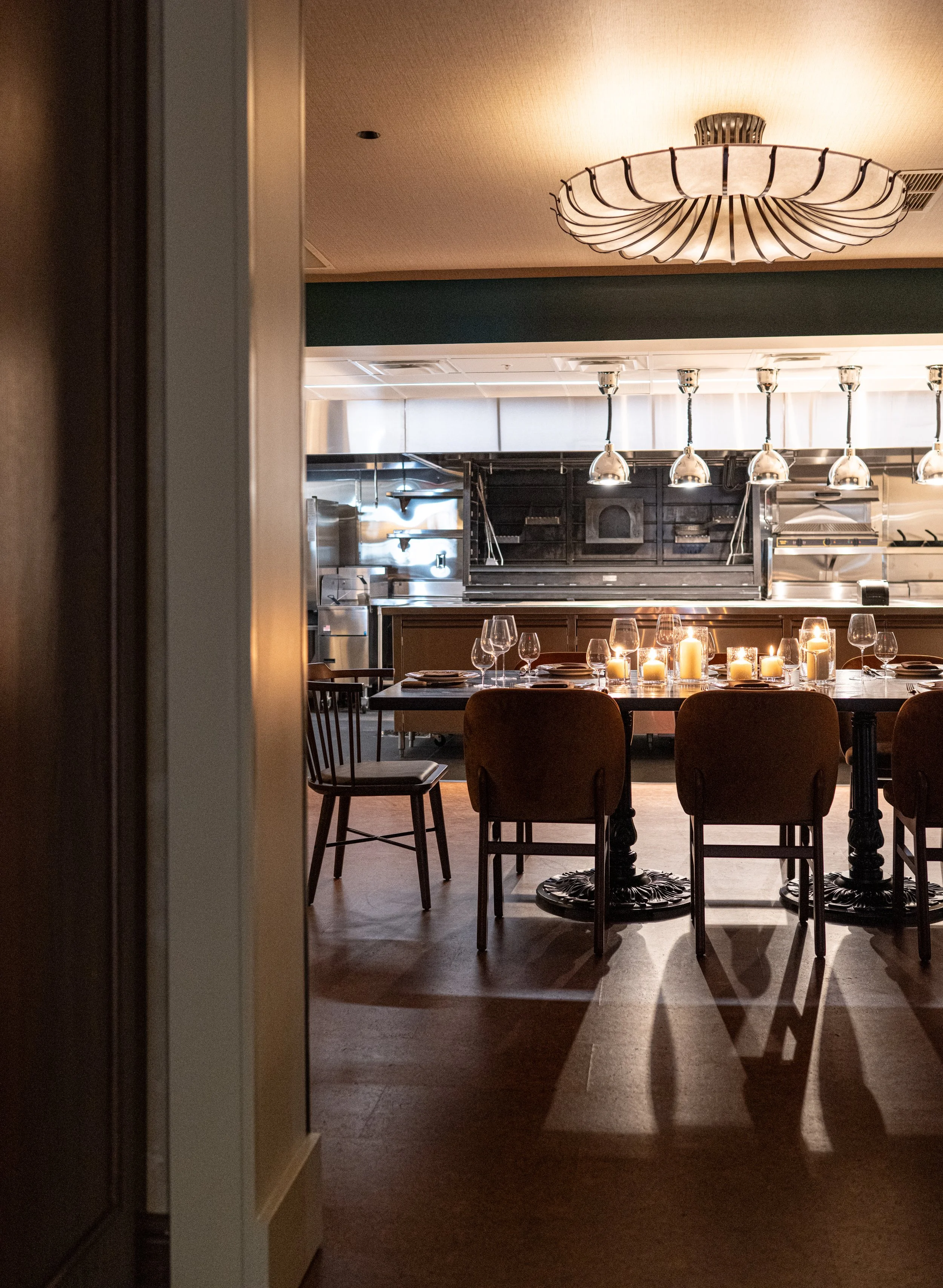 A dining room set up with a long table decorated with candles and wine glasses, chairs, and a chandelier hanging from the ceiling. The background shows a kitchen area with stainless steel appliances and lighting.