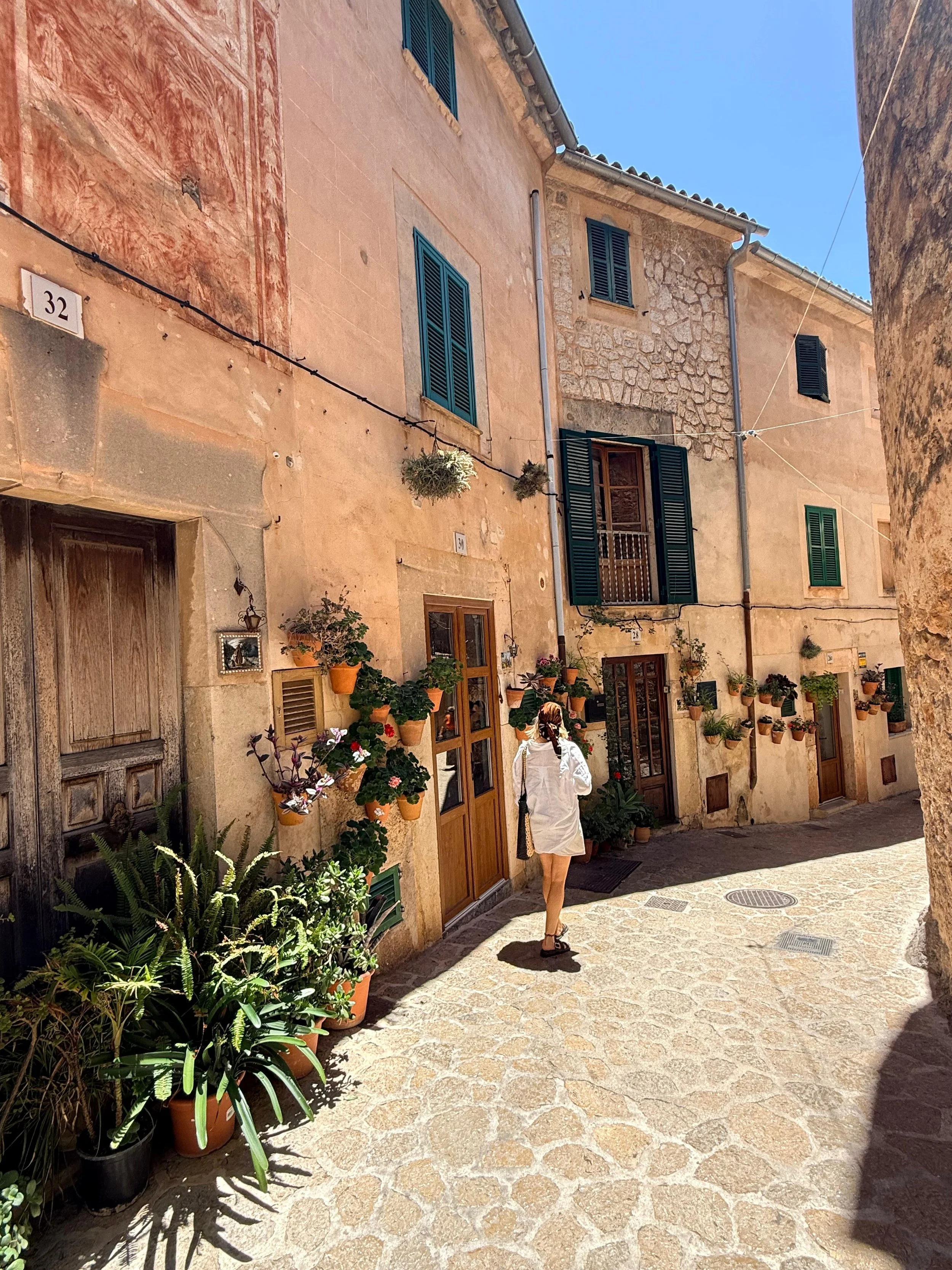 A woman with long hair, wearing a white dress, walking along a cobblestone street next to a row of potted plants and rustic buildings with blue shutters.