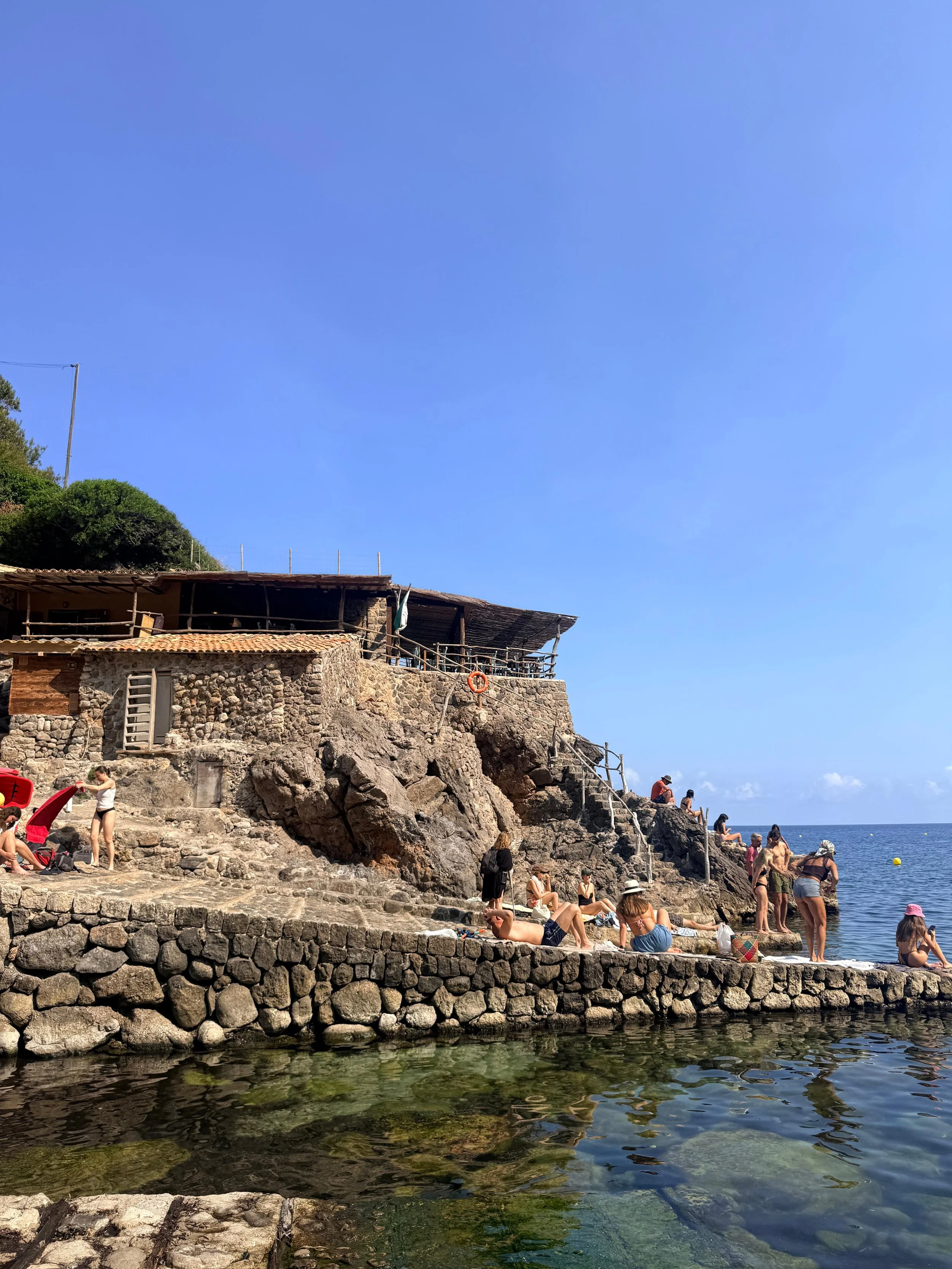 People relaxing and swimming on rocky steps along the shore of a sunny beach with a blue sky and ocean in the background.