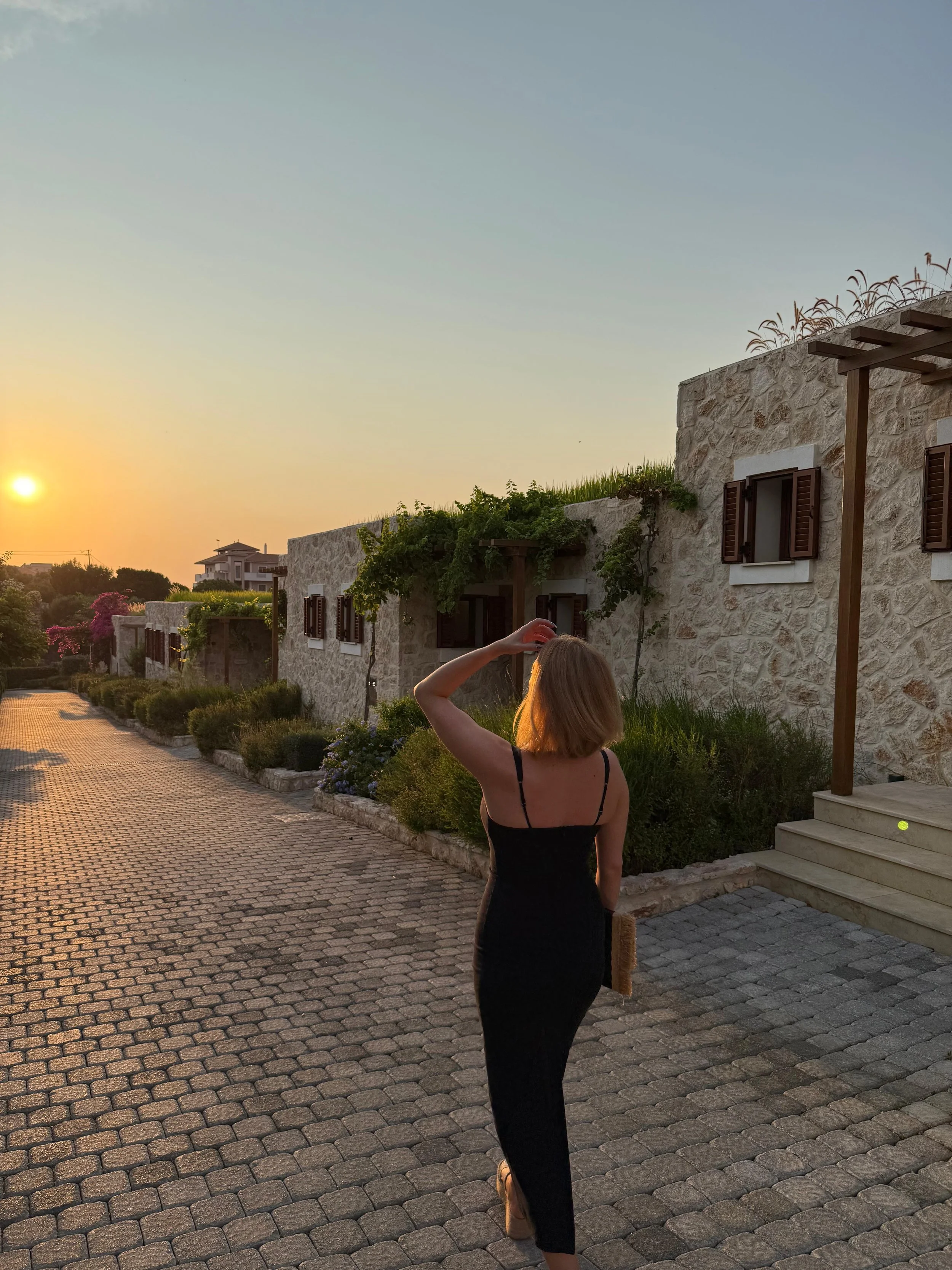 A woman in a black dress standing on a cobblestone path, holding her head with her right hand, during sunset outside a stone building with windows and greenery.