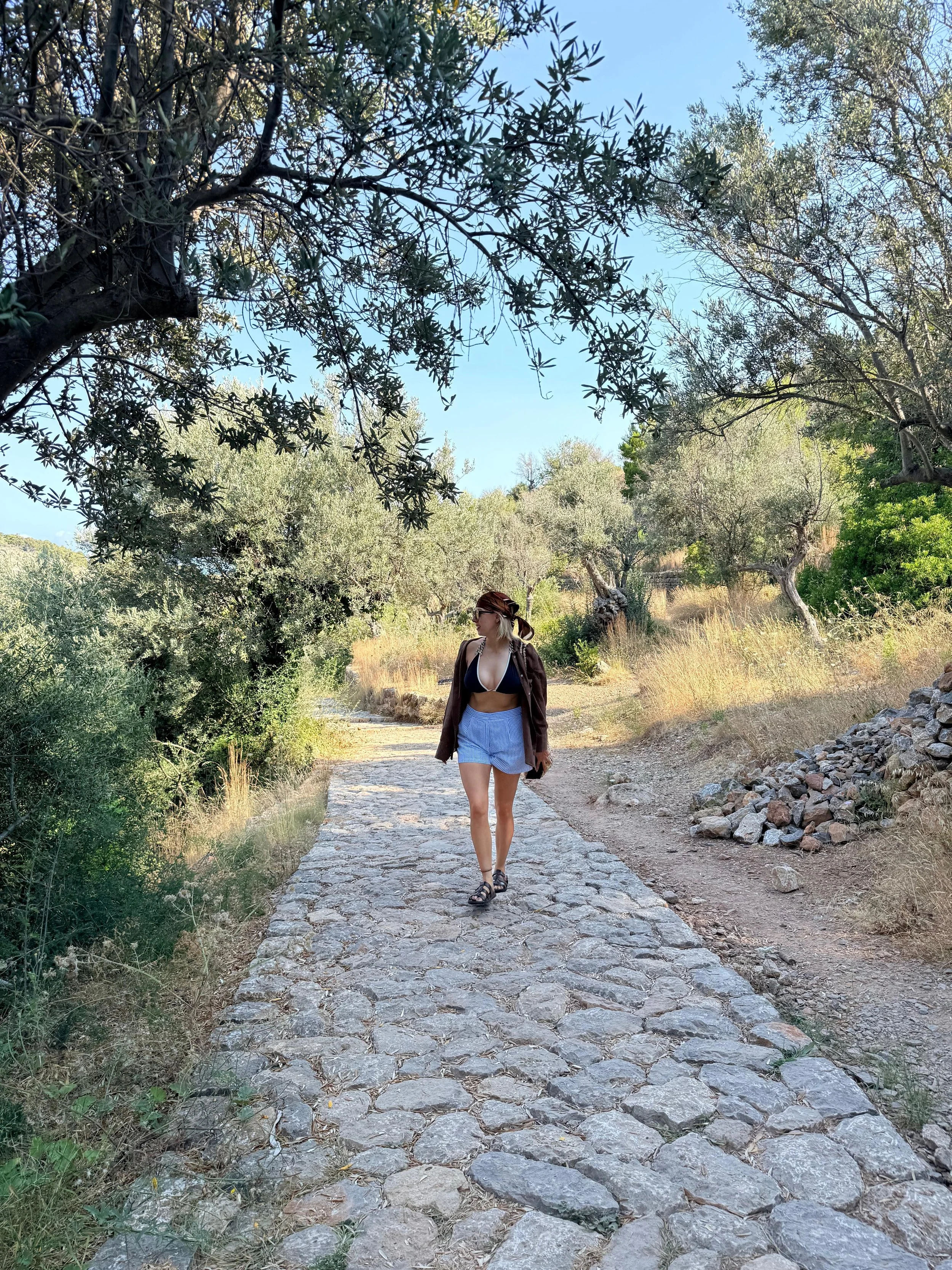 A woman walking on a stone path through a grassy and wooded area with trees and blue sky overhead.