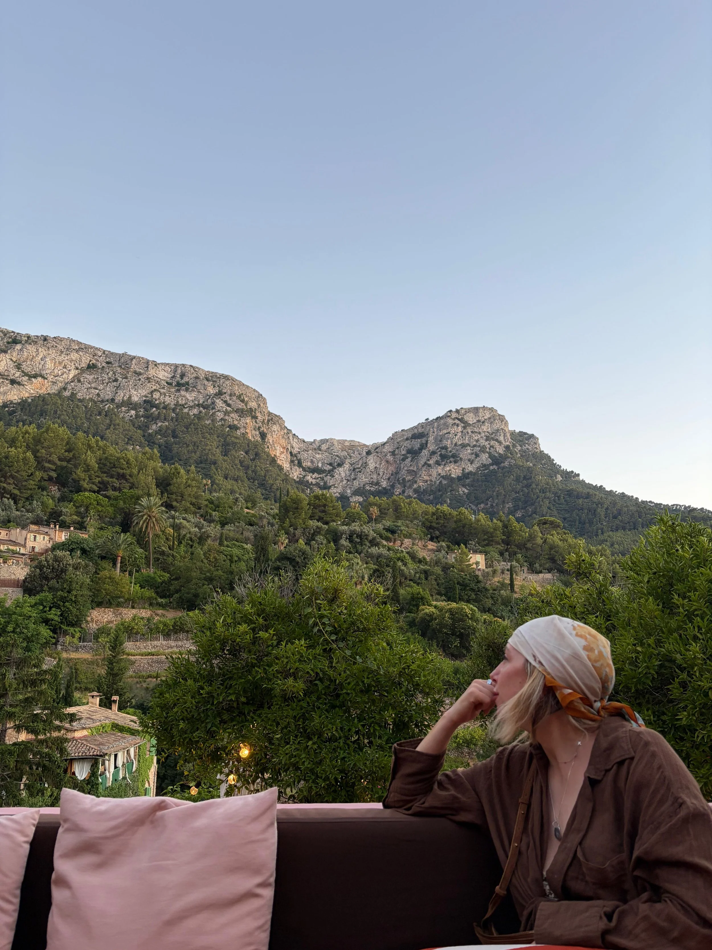 A woman with blonde hair wearing a beige headscarf and brown jacket sits outdoors, resting her chin on her hand, with a scenic mountain landscape and green trees in the background.