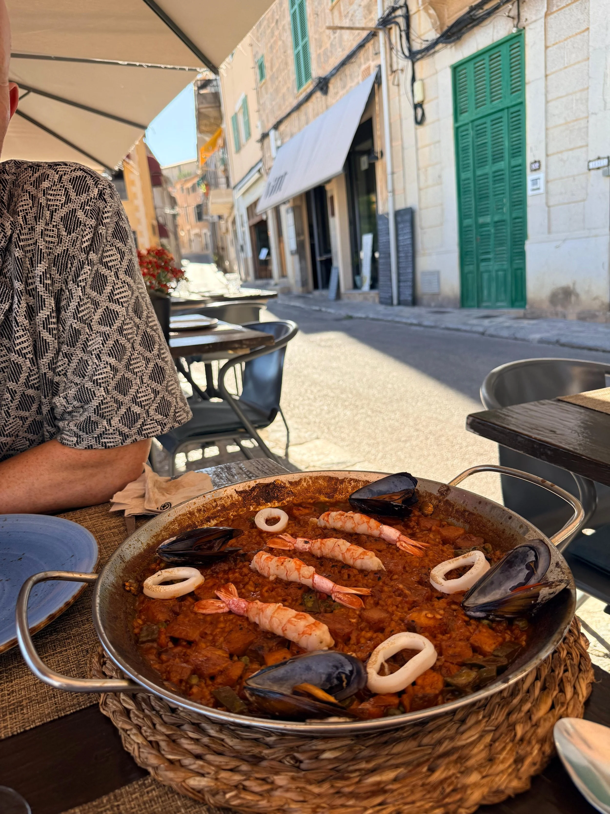 A seafood dish in a large, shallow pan with shrimp, squid rings, mussels, and other seafood in a tomato-based sauce, on a table at an outdoor restaurant.