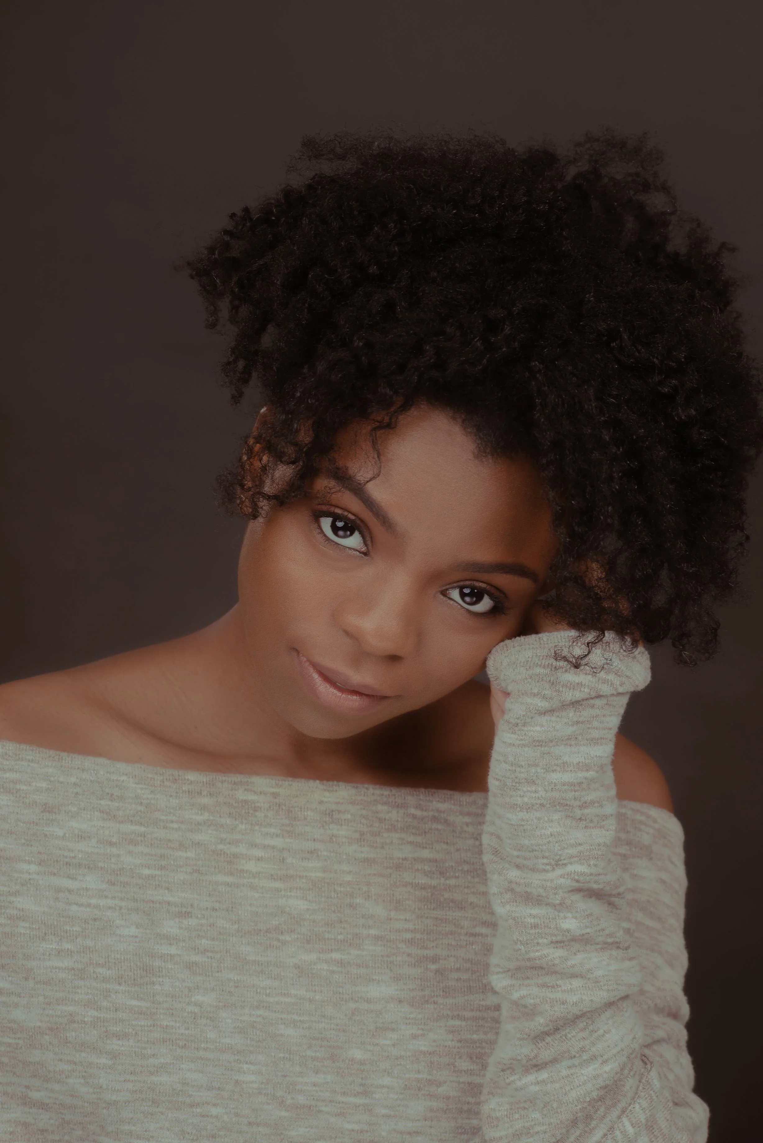 A young, Black woman with beautiful curly hair, resting her face on her hand, looking into the camera, wearing a blush off-the-shoulder sweater against a dark background.