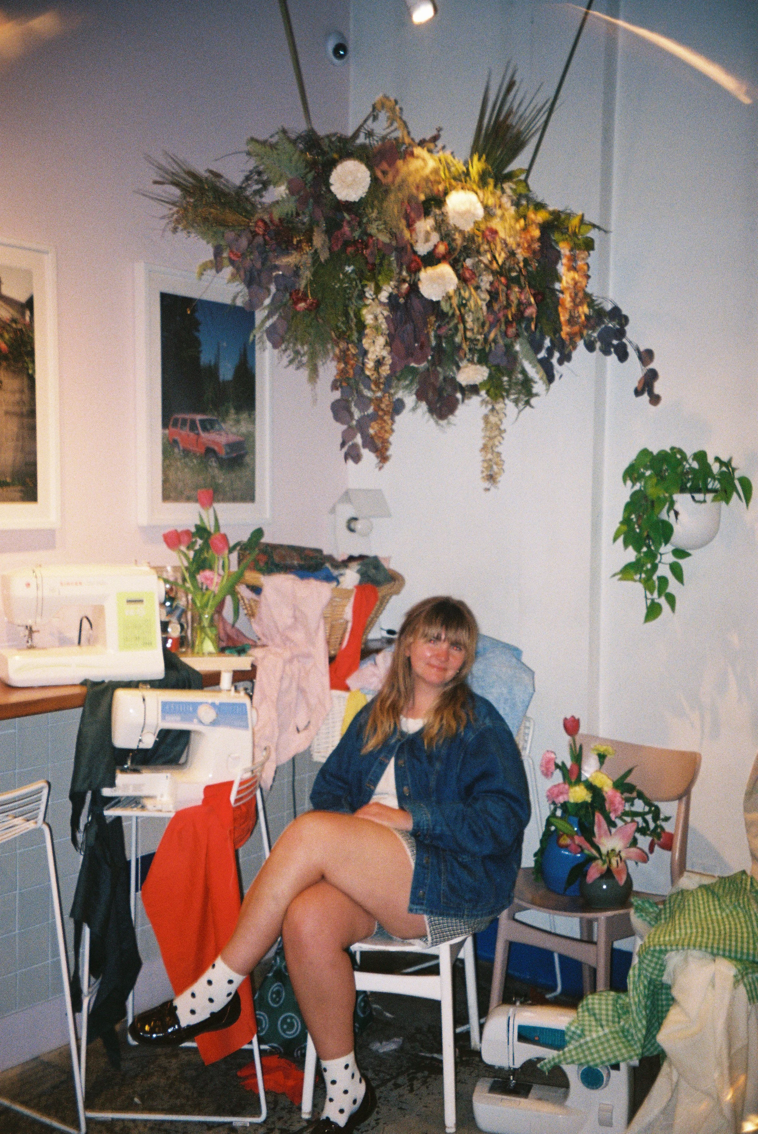 A woman sitting on a chair with floral arrangements and sewing machines in a room with artwork on the walls. She is wearing a denim jacket, shorts, and polka dot socks, smiling at the camera.