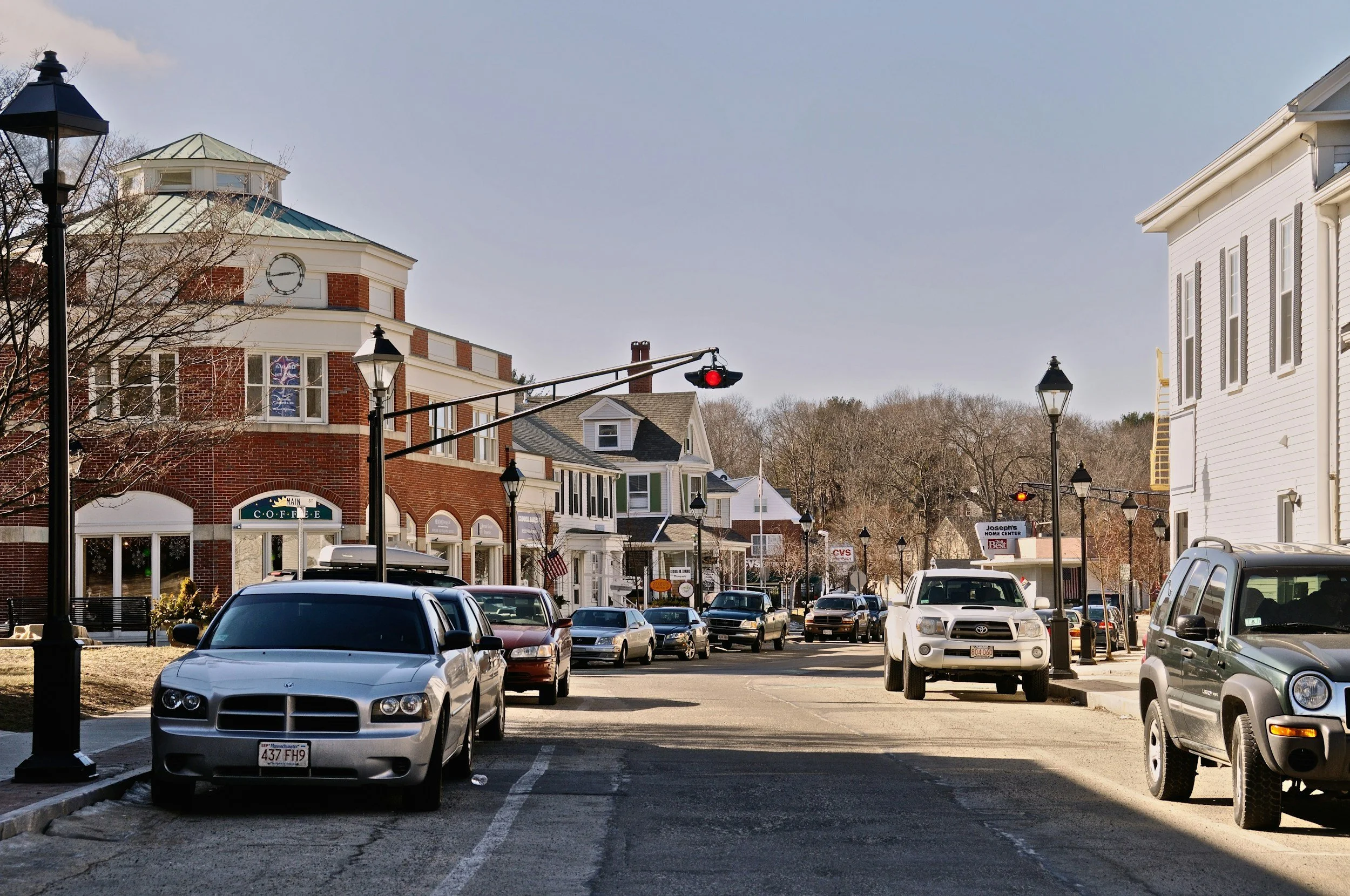 A small town street with parked cars, decorative street lamps, and a mix of brick and white buildings, including a coffee shop and other storefronts, during daylight.
