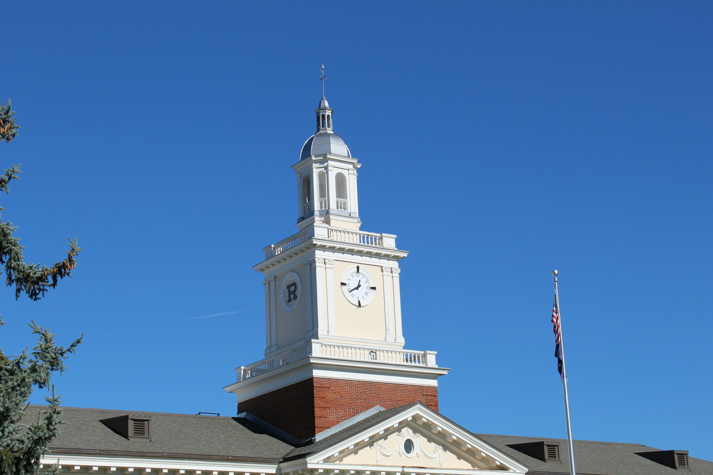 A white clock tower with a wagon wheel clock, on top of a brick building, with an American flag on a flagpole in front, against a clear blue sky.