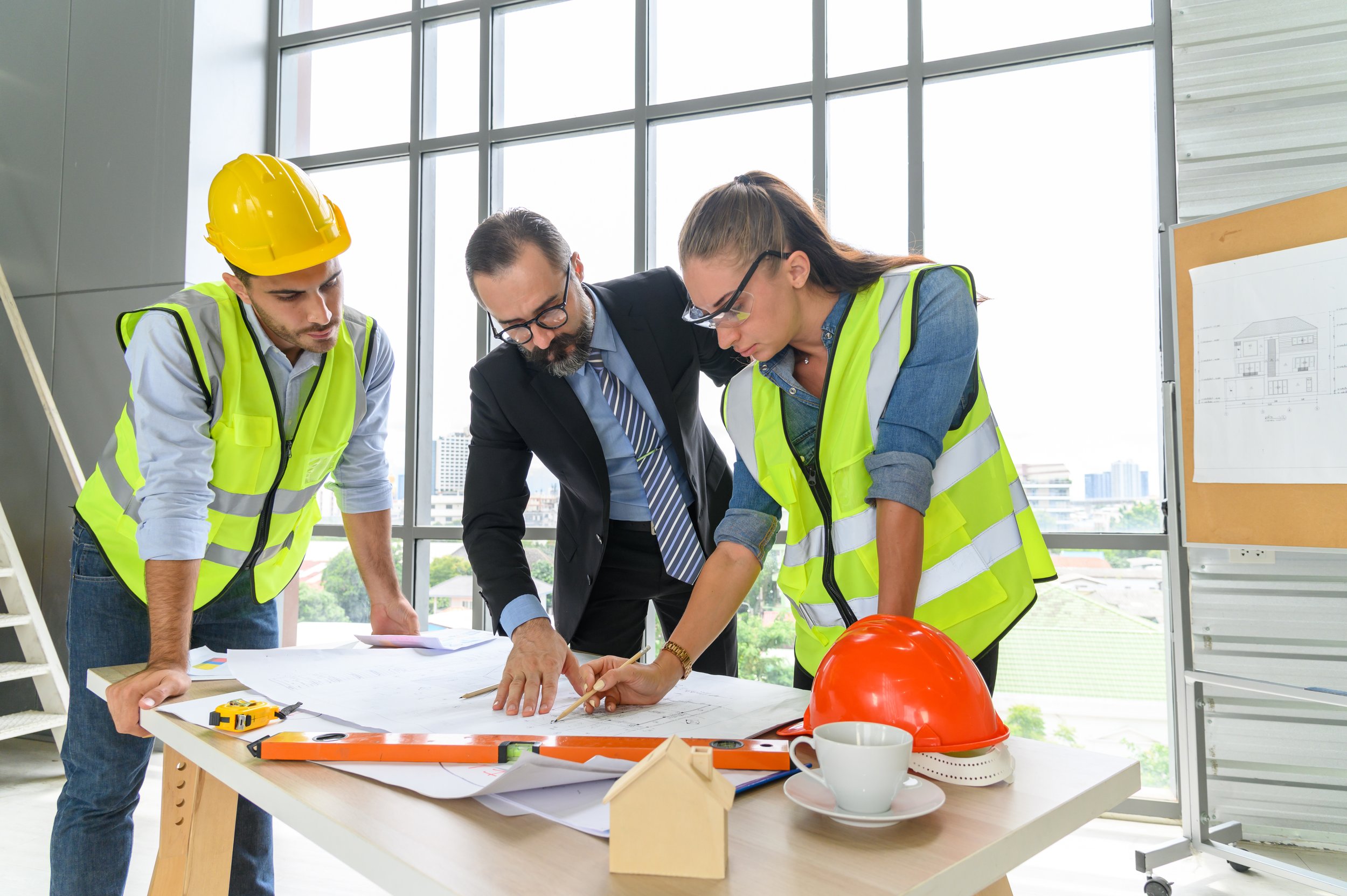 Three construction professionals, two in safety vests and one in a suit, reviewing blueprints at a table in a modern office with large windows.